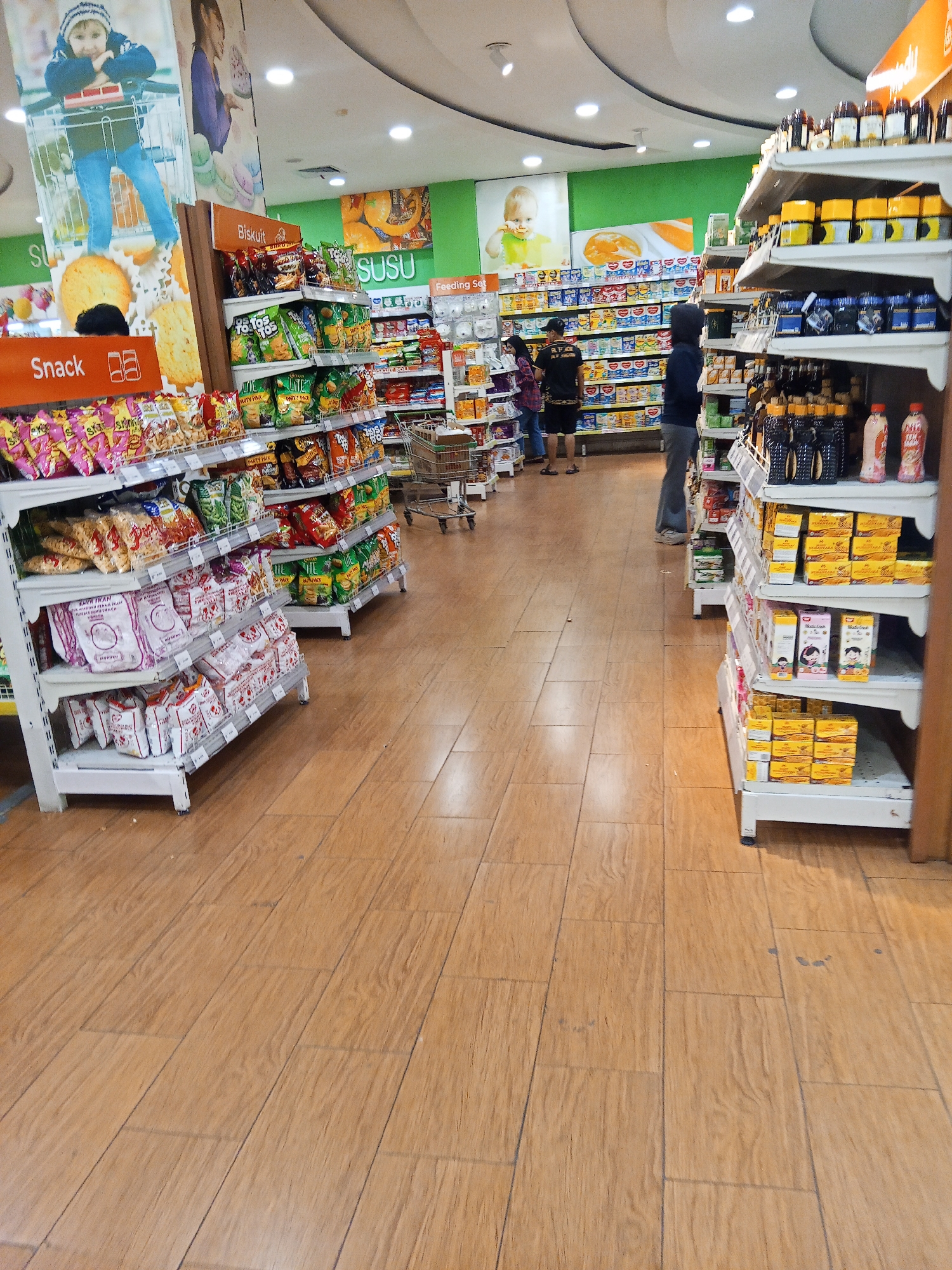 The image depicts the interior of a brightly lit supermarket, likely located in Lubuklinggau, Indonesia, with shelves stocked with various food items and beverages. In the foreground, a wooden floor with a tiled pattern extends throughout the aisle. To the left, shelves are filled with colorful snack bags and packaged goods, including "Snack" and "Biskuits" branding visible. A large banner featuring a child in a shopping cart is prominently displayed on the left wall, along with other promotional visuals.
In the middle ground, more shelving units display items such as "SUSU" (milk) products, baby food, and various packaged goods. Several shoppers are visible, including a person wearing a black t-shirt and cap, another in a dark hoodie, and two individuals further down the aisle. A shopping cart is positioned in the aisle, indicating active commerce.
To the right, a tall shelving unit is densely packed with bottled products, including what appear to be sauces and condiments, as well as boxes of other goods. The overall environment suggests a well-organized retail space designed for grocery shopping, with an emphasis on variety and accessibility of products. The lighting is even, illuminating the merchandise and creating a welcoming atmosphere.