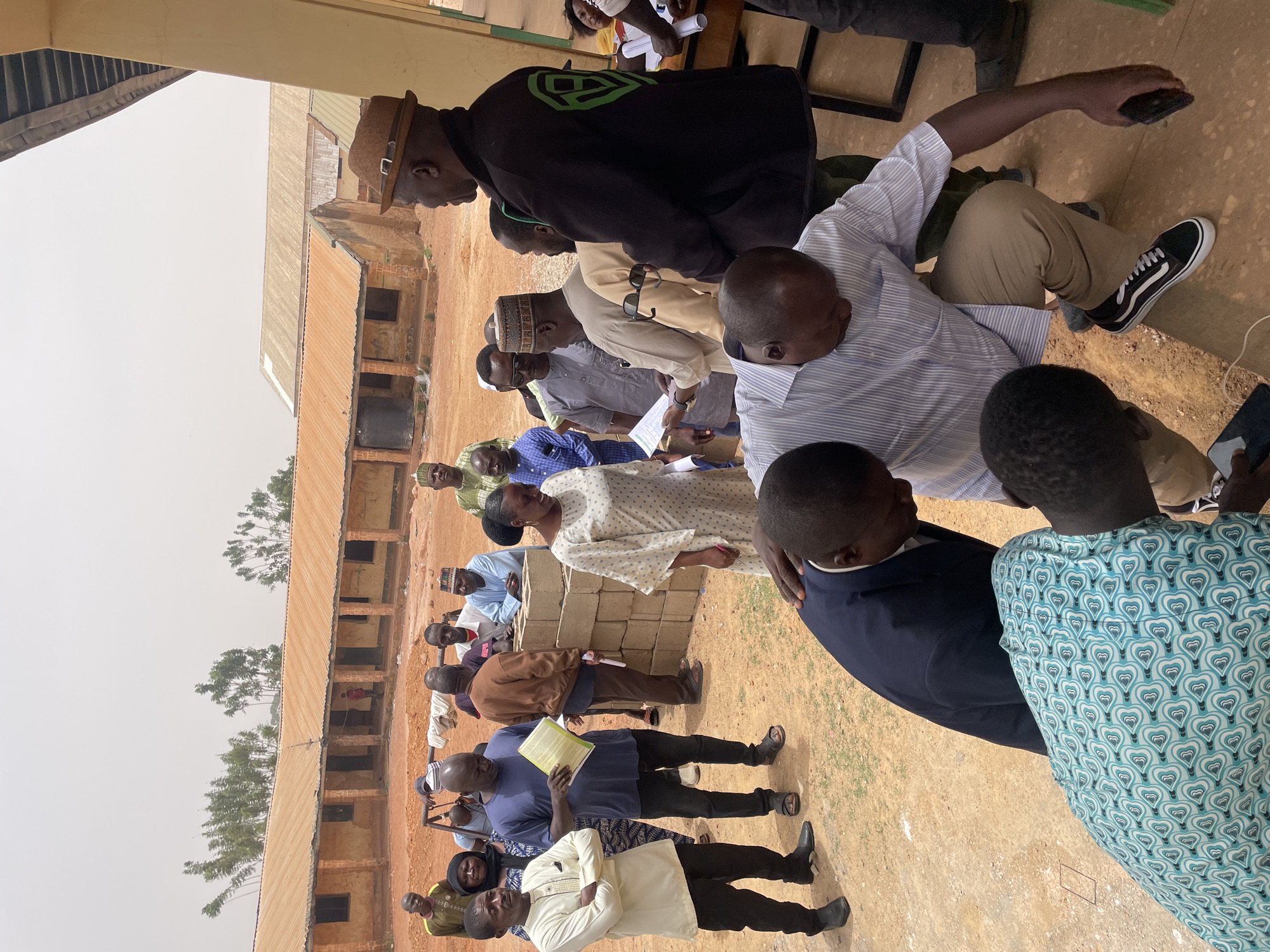 A group of people are gathered outdoors, presumably in Bukuru, Nigeria, as indicated in the prompt. The setting appears to be an open area with a building in the background. The building is a long, low structure with multiple windows. The ground is sandy or dirt-covered.
The group consists of men and one woman, most are dressed in casual attire, although some are wearing suits. A man in the background of the group is wearing a hat. Some of the people are standing, while others are sitting on benches and cinder blocks. They seem to be engaged in a meeting or discussion, as many are holding papers and looking at each other. The picture seems to have been taken at mid-day.