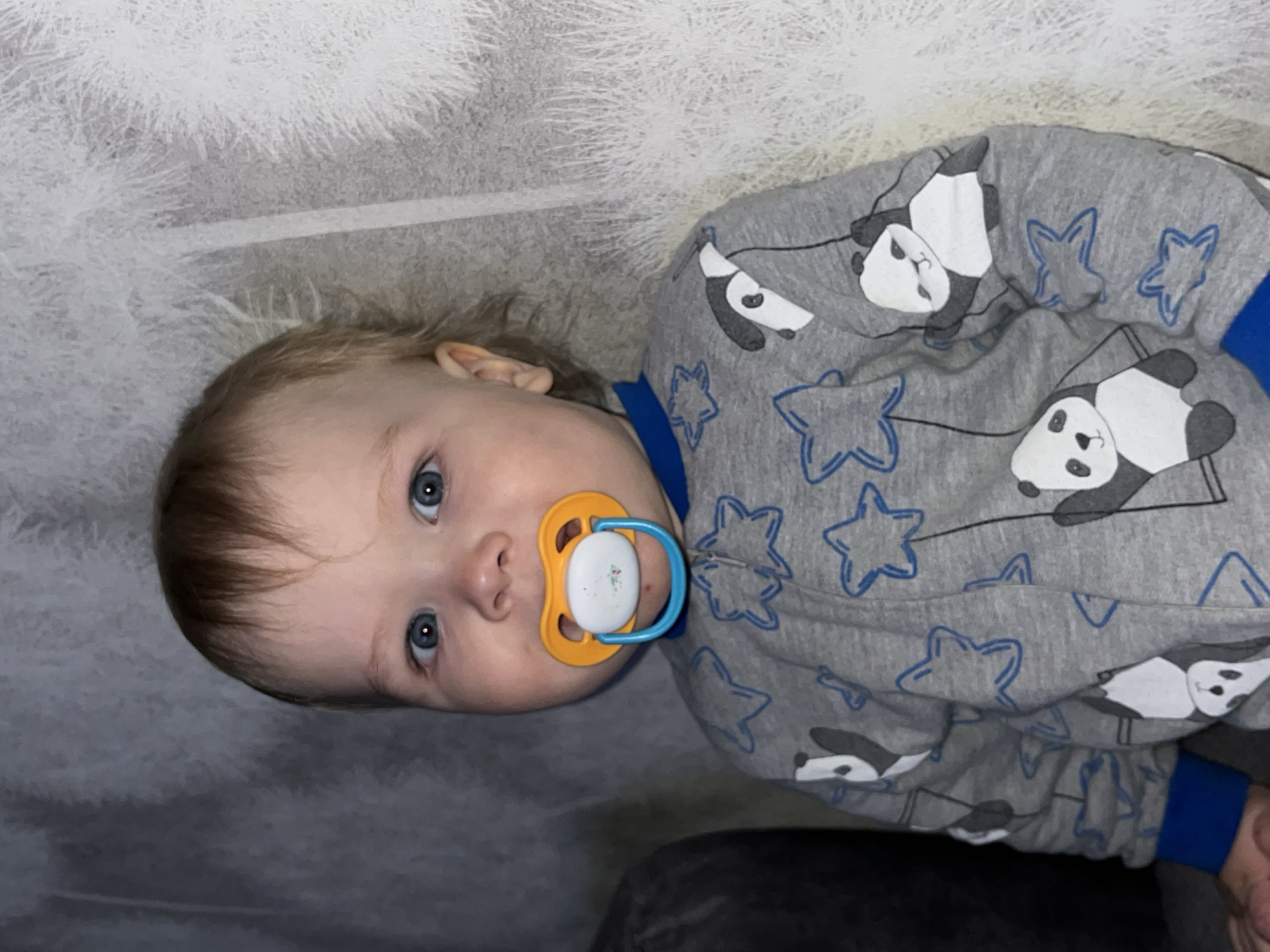 A young child, estimated to be between 1 and 2 years old, is lying on a surface. The child has fair skin, light brown hair, and blue eyes, and a pacifier in their mouth. The child is wearing gray pajamas patterned with blue stars and images of pandas. The background appears to be a gray surface, possibly a blanket or bedding. The exact location within Chaikovsky, Russia, is not discernible from the image alone.