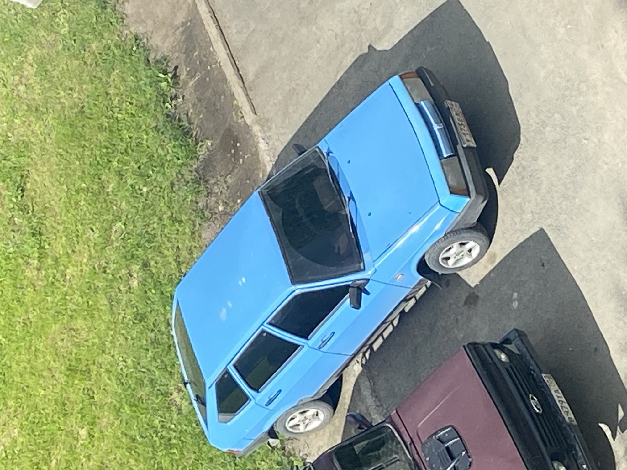 The image shows two parked cars on a paved surface next to a grassy area. The primary subject is a bright blue, five-door hatchback, viewed from a high angle. The car has tinted windows, silver alloy wheels, and a license plate visible on the rear. To the left and slightly in front of the blue car is the front section of a darker colored vehicle, possibly a maroon SUV or sedan. This second vehicle also has tinted windows and a license plate. The environment appears to be an outdoor urban setting, potentially a parking lot or residential area, with a patch of green grass to the upper left and a concrete or asphalt ground surface. The sun is casting shadows from the cars onto the ground. The overall impression is a still, everyday scene.