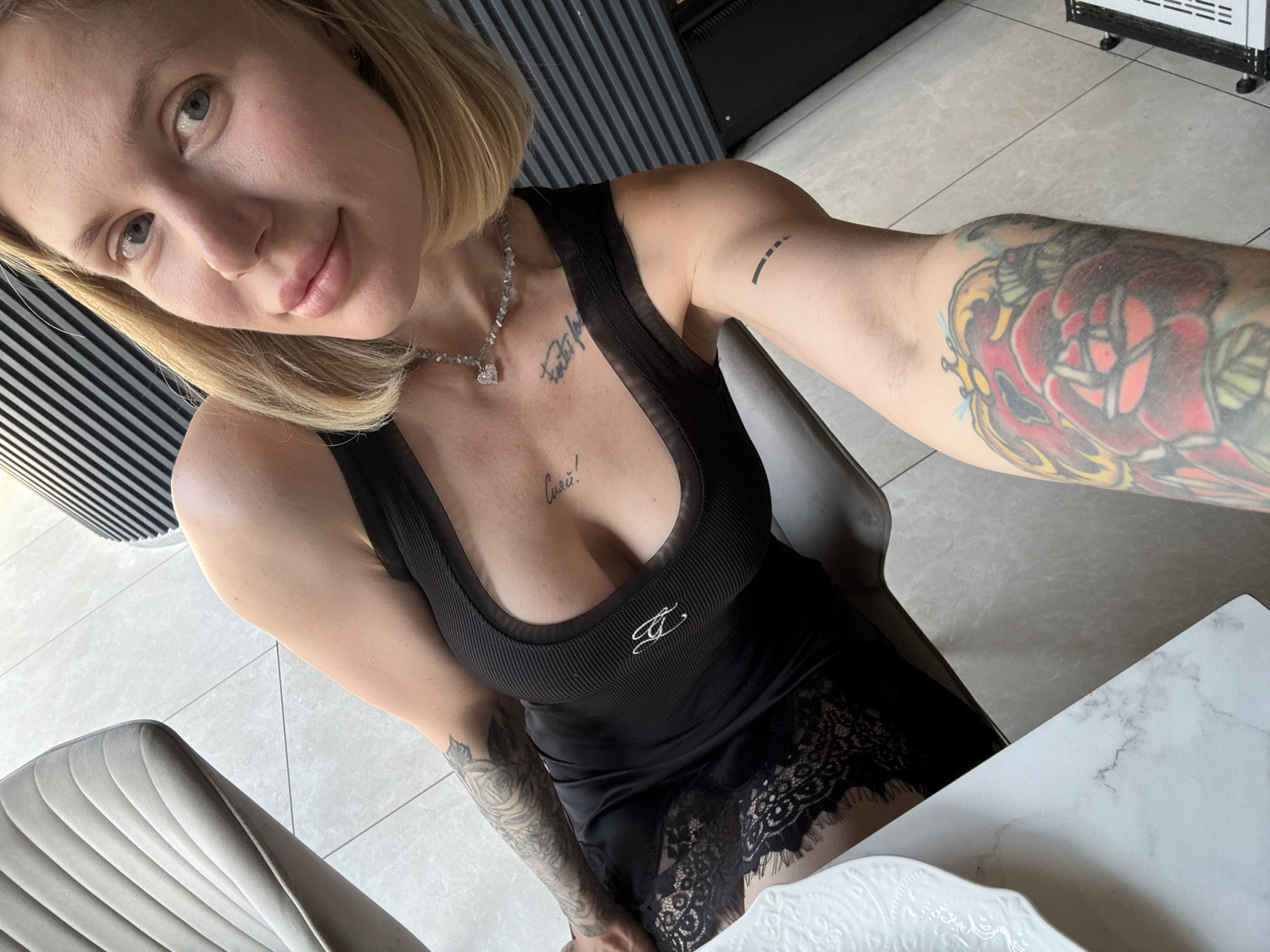 In Ust'-Muny, Russia, a woman with blonde hair and tattoos is taking a selfie. She is wearing a black dress with lace trim and a necklace. She is sitting in a chair in front of a table. The room has tiled floors and a wall with black and white striped panels.
