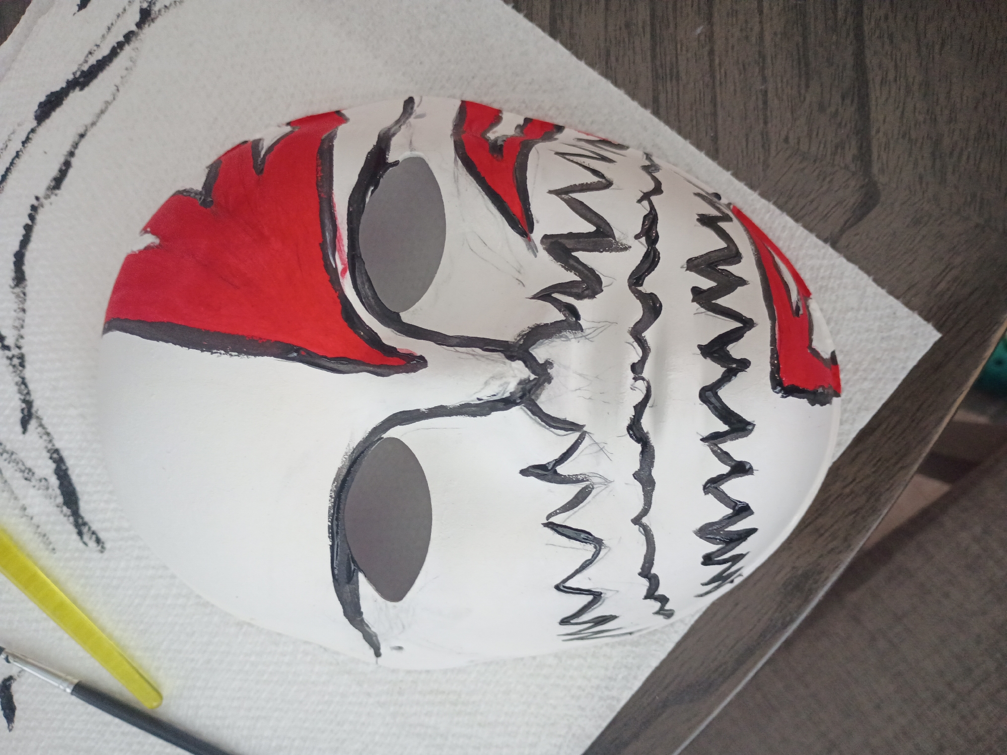 A white mask with black and red paint is resting on a white paper towel. The mask has eye holes and a design featuring red shapes at the top and black jagged lines down the sides. A paintbrush and a yellow tool are visible to the left of the mask. The surface beneath the paper towel appears to be wooden. The location of this image is unknown and cannot be determined to be in Los Angeles, United States of America based on its content.