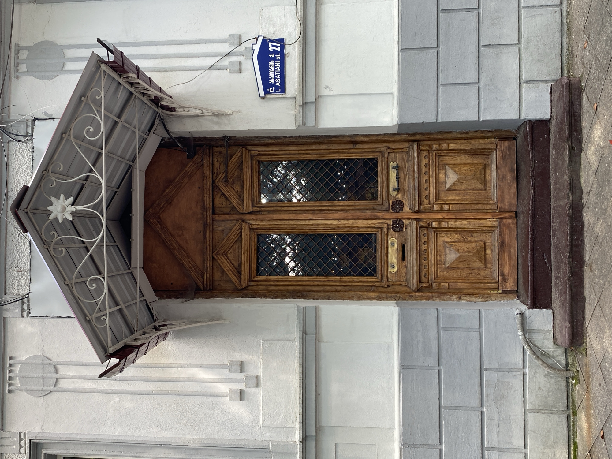 The image shows a close-up of a double wooden door in Batumi, Georgia. The door is elaborately carved and features two windows with diamond-patterned grates. Above the door is a decorative metal awning with scrollwork and a floral motif. The door is set into a white, stucco-covered building with gray brick detailing at the base. A blue and white street sign with Georgian text and the number "27" is visible on the wall to the right of the door. A flexible gray pipe runs from the wall to the ground near the steps leading up to the door. The overall scene depicts an architectural detail of an older building in an urban setting.