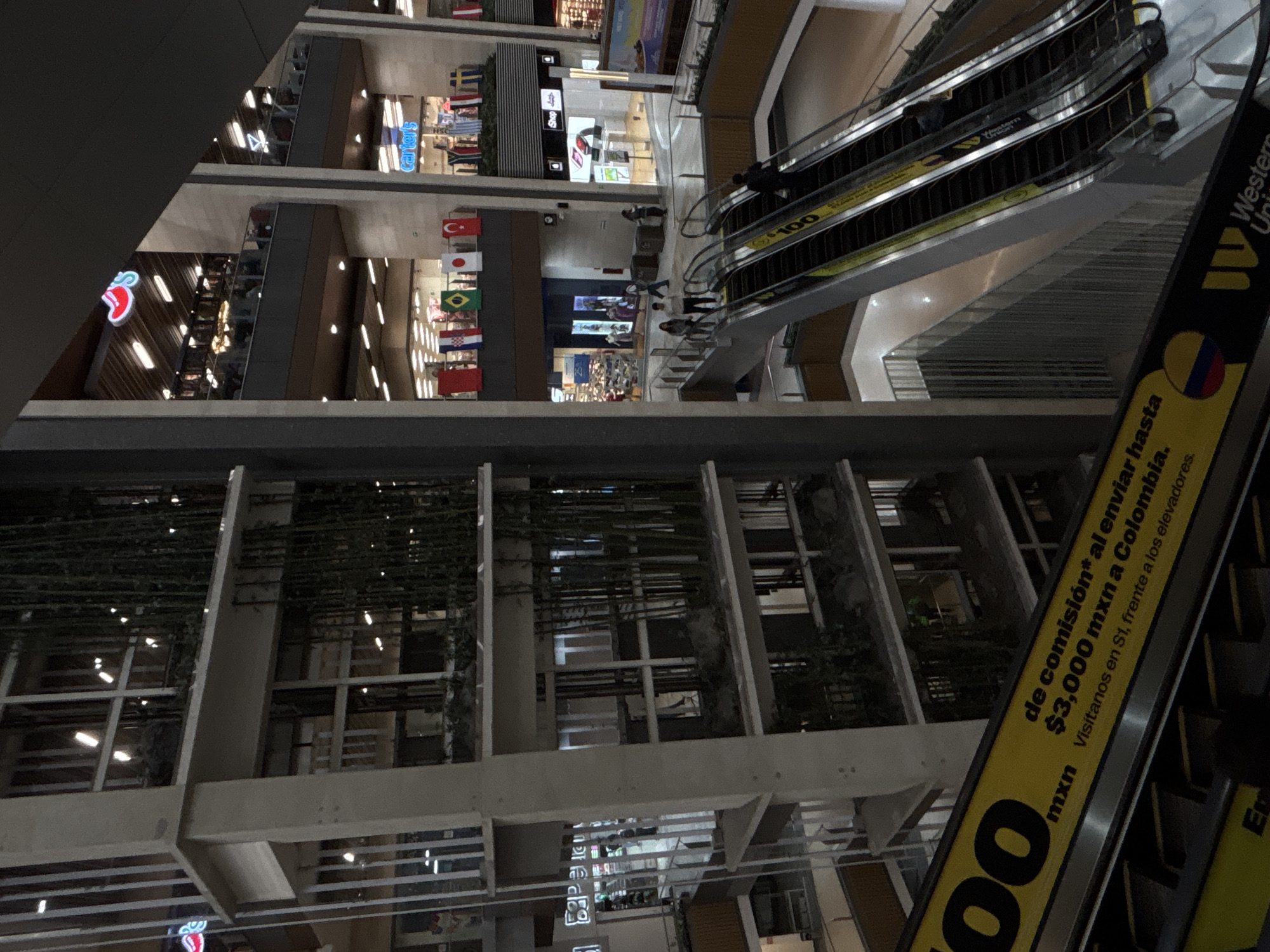 The image depicts the interior of a multi-level shopping mall, likely in Mexico City, Mexico, as indicated by the presence of the Colombian flag and the text "mxn" (Mexican Peso) on an escalator banner. Multiple escalators ascend through the building, with people visible using them. On various levels, shops are discernible, some with illuminated signs. One prominent banner on an escalator reads "de comisión* al enviar hasta $3,000 mxn a Colombia. Visítanos en S1, frente a los elevadores." This translates to "commission* on sending up to $3,000 mxn to Colombia. Visit us on S1, in front of the elevators." This suggests a money transfer service is advertised. Flags of various countries are displayed on one of the upper levels, possibly for a promotional event. The architecture features concrete columns and balconies with vertical elements, and some areas have hanging plants or bamboo-like structures. The lighting appears to be artificial and interior.