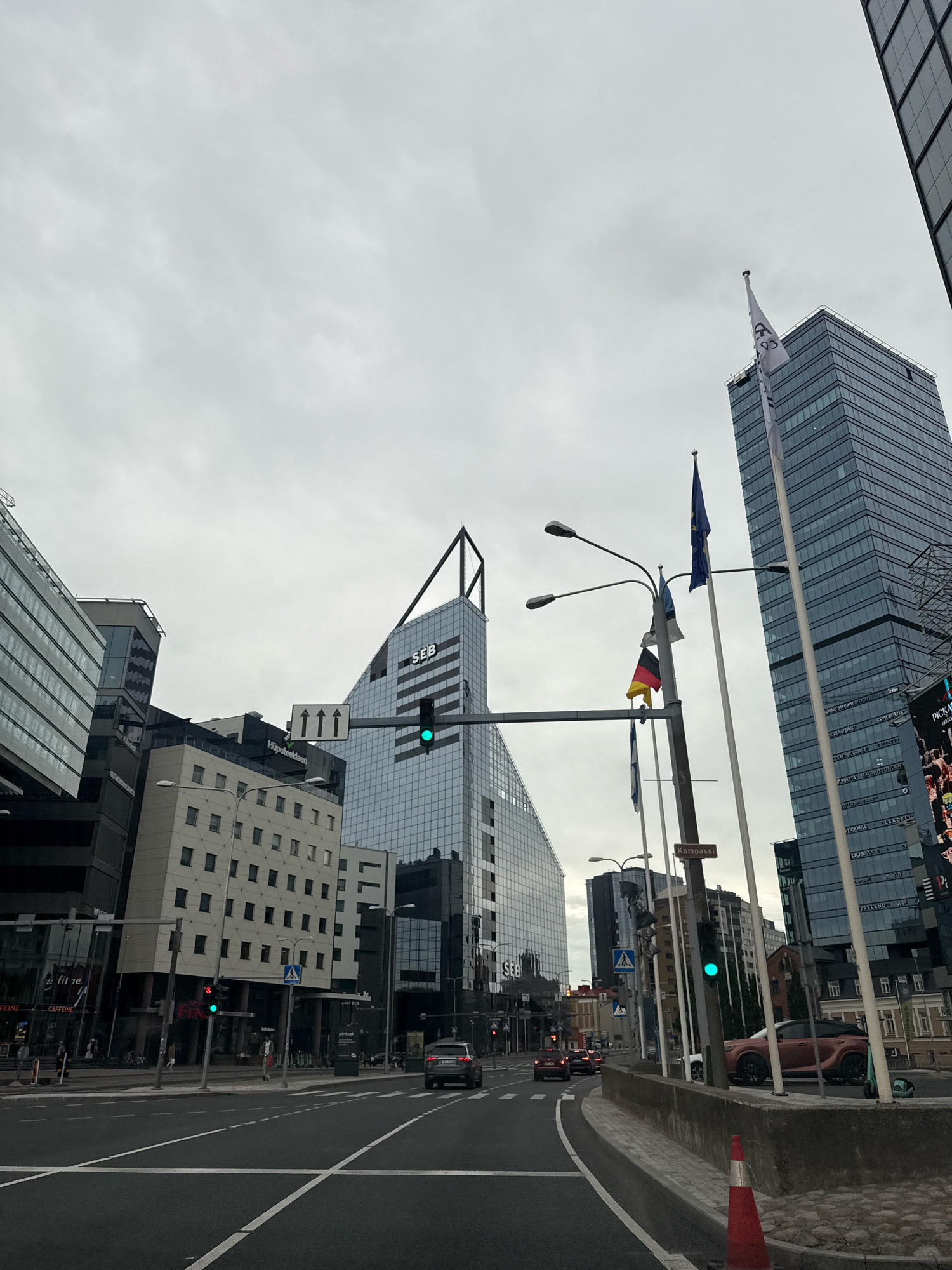 The image depicts a street view in Tallinn, Estonia, specifically in the business district. Modern glass-fronted buildings dominate the skyline, including the SEB headquarters. The sky is overcast. Traffic lights are visible, with a green light illuminated. Several cars are on the road, and a few pedestrians can be seen on the sidewalks. Flags, including the European Union flag, the Estonian flag, and the German flag, are displayed on flagpoles. A traffic cone is present on the right side of the road. The architecture suggests a contemporary urban environment.