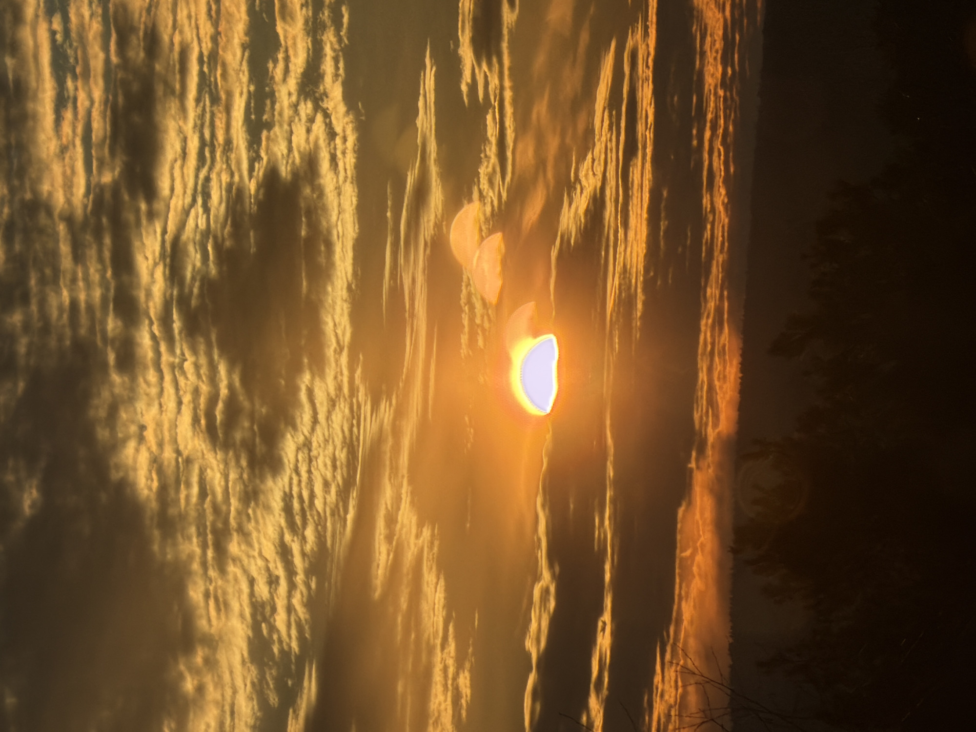 The image shows a sunset over a landscape in Chaikovsky, Russia. The sky is filled with streaky, golden-colored clouds, likely reflecting the setting sun. The sun itself is visible through the clouds, appearing as a bright, golden orb with a hazy glow. To the right, there is a darker area possibly indicating trees or a forested area. The overall scene suggests a tranquil, natural setting bathed in the warm light of the setting sun.