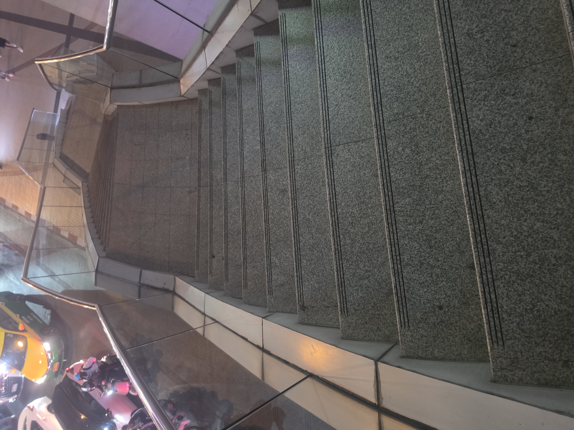 An elevated, downward-looking shot captures a wide, curved outdoor staircase leading to a busy street below. The steps are made of speckled grey granite or stone, each tread featuring multiple parallel dark non-slip grooves. A modern railing system, constructed from clear glass panels and metallic frames, follows the curve of the staircase on the left side.
The highly reflective surfaces of the glass and metal railing capture numerous reflections, showcasing distorted images of the street activity, various lights, and indistinct shapes of people. At the bottom left, the street level is visible with artificial lighting. Vehicles are present, including a characteristic yellow and green taxi, a motorbike with its headlamp illuminated, and parts of another white vehicle.
The scene appears to be set at dusk or night, indicated by the reliance on artificial light sources and the illumination from vehicle head/taillights. The overall atmosphere is urban and active. The architecture suggests an elevated pedestrian walkway or access to a public transport system, common in bustling cities. The prominent yellow and green taxi is a strong contextual clue for Bangkok, Thailand. No discernible text is visible.