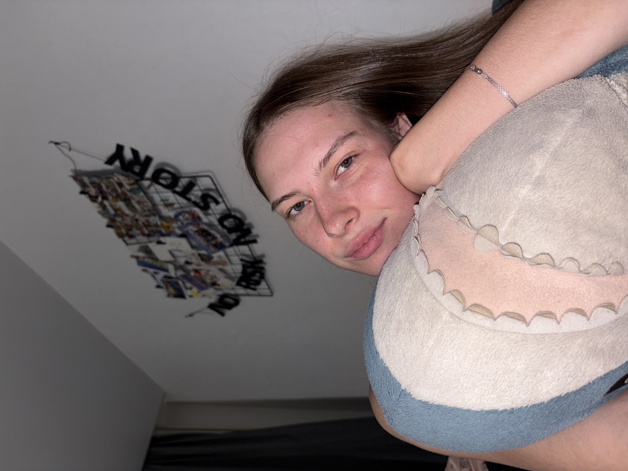 A young Caucasian woman with long brown hair and blue eyes is lying down, resting her head on a plush shark toy. She is wearing a light-colored top. Her skin appears flushed, and she has minimal makeup on. Behind her, a white wall displays a wire grid with several photos attached and black lettering that reads "NO RISK NO STORY." A dark curtain is visible to the left of the frame. The scene is indoors, likely a bedroom, and the lighting suggests it is nighttime. There is no discernible information in the image to determine its location in the city of Perm, Russia.