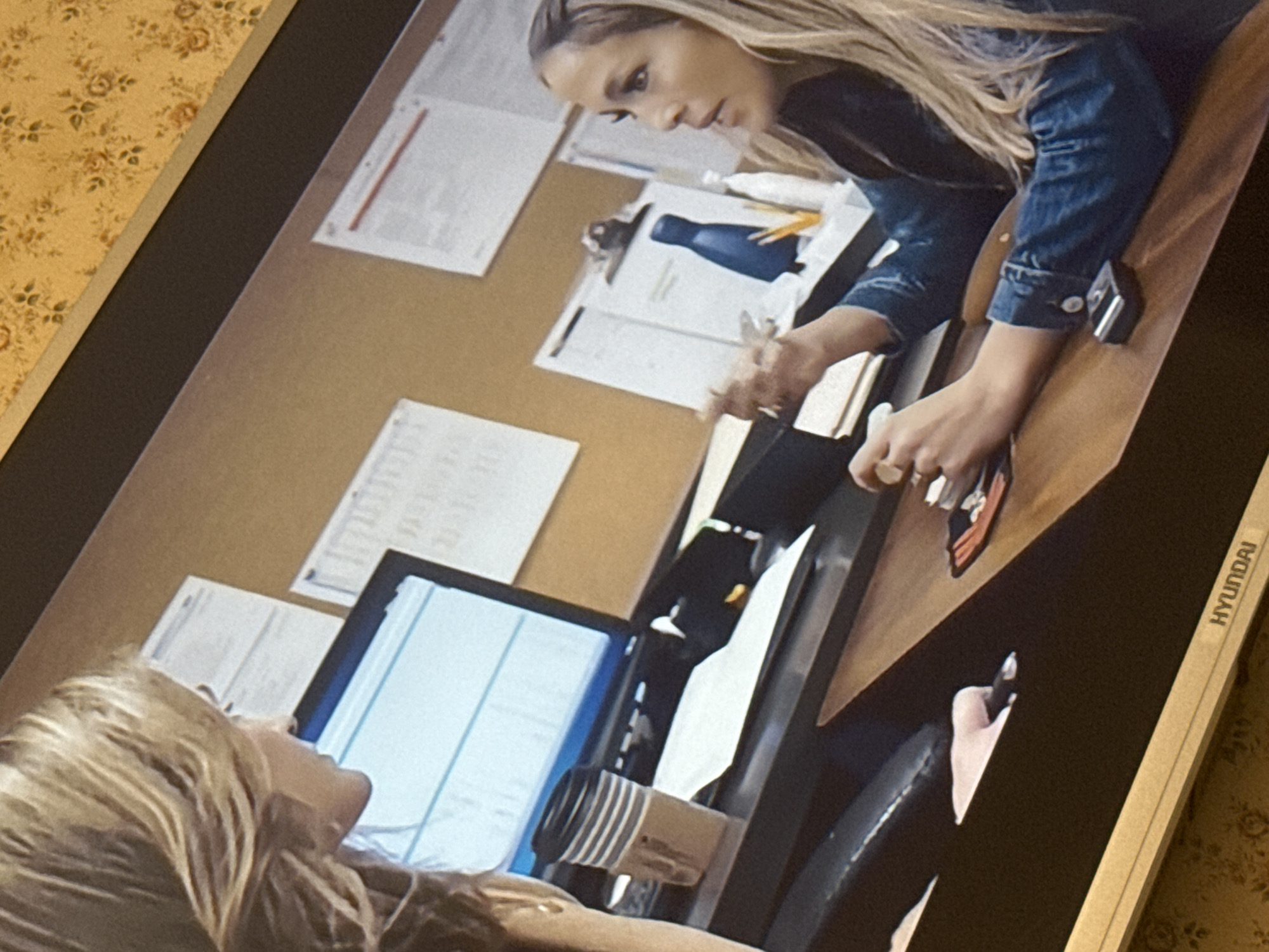 The image shows a scene from what appears to be a television or computer screen. Two women with blonde hair are visible in the frame. One woman is leaning over a desk, wearing a denim jacket, and interacting with objects on the desk. The desk appears to be covered with papers, a blue water bottle, and other office items. The other woman is sitting in what appears to be a vehicle, partially visible in the lower part of the frame, with a computer monitor in front of her. The brand "HYUNDAI" is visible on the side of the monitor. The background behind the desk appears to be a cork board with more papers attached. The overall environment seems to be an office setting. There is no information given or visible in the image that would indicate the location as Mytishchi, Russia.