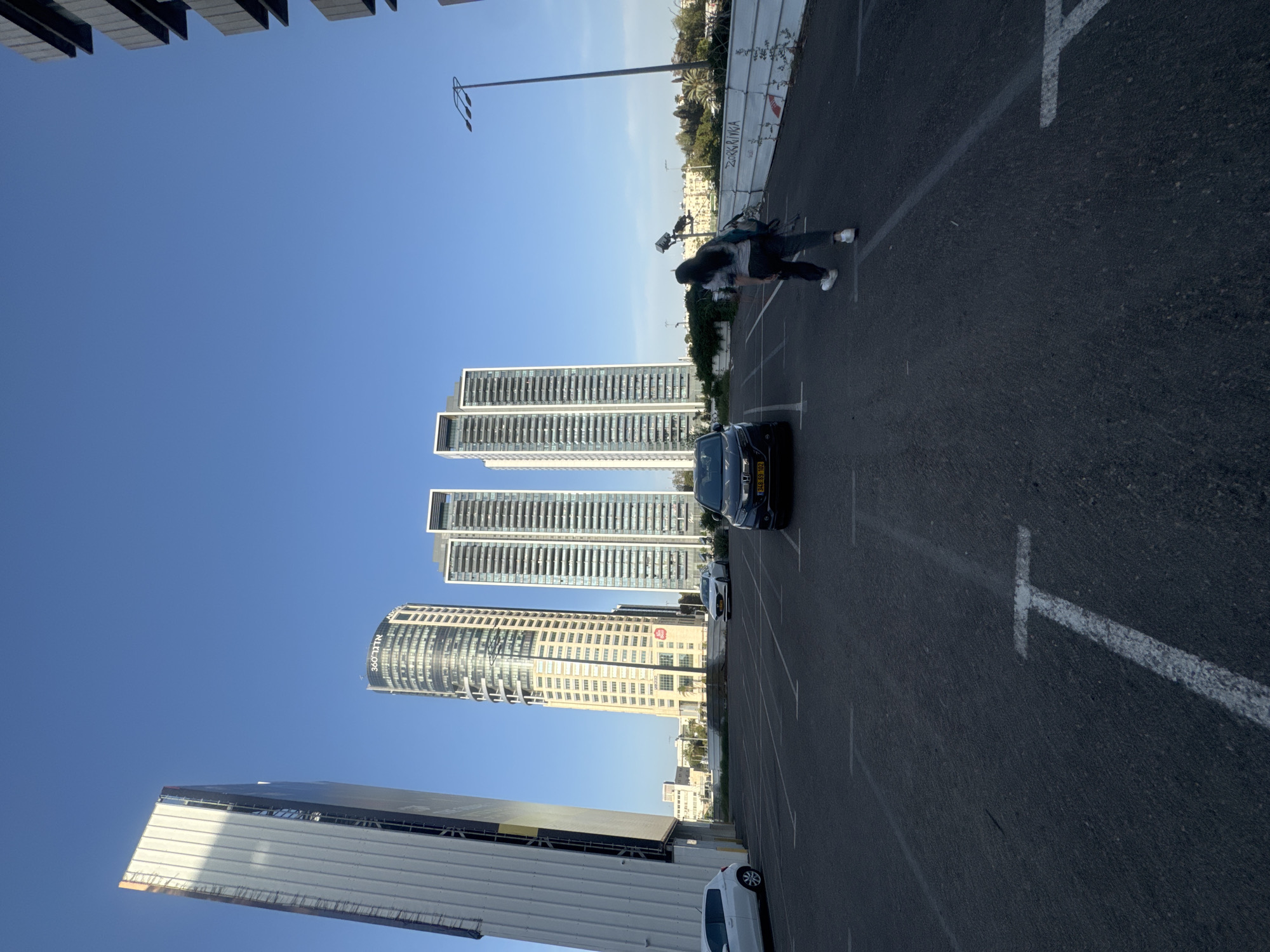 The image captures an outdoor scene in Tel Aviv, Israel. Four tall buildings dominate the left side of the frame, under a clear blue sky. The building closest to the bottom displays the text "אדגר 360". A large white truck is situated along the bottom edge. A black car is parked mid-ground. In the upper-right, a person is seen walking, their shadow cast on the pavement. The ground is marked with parking lines, indicating a parking lot. A tall streetlight stands next to a fence.