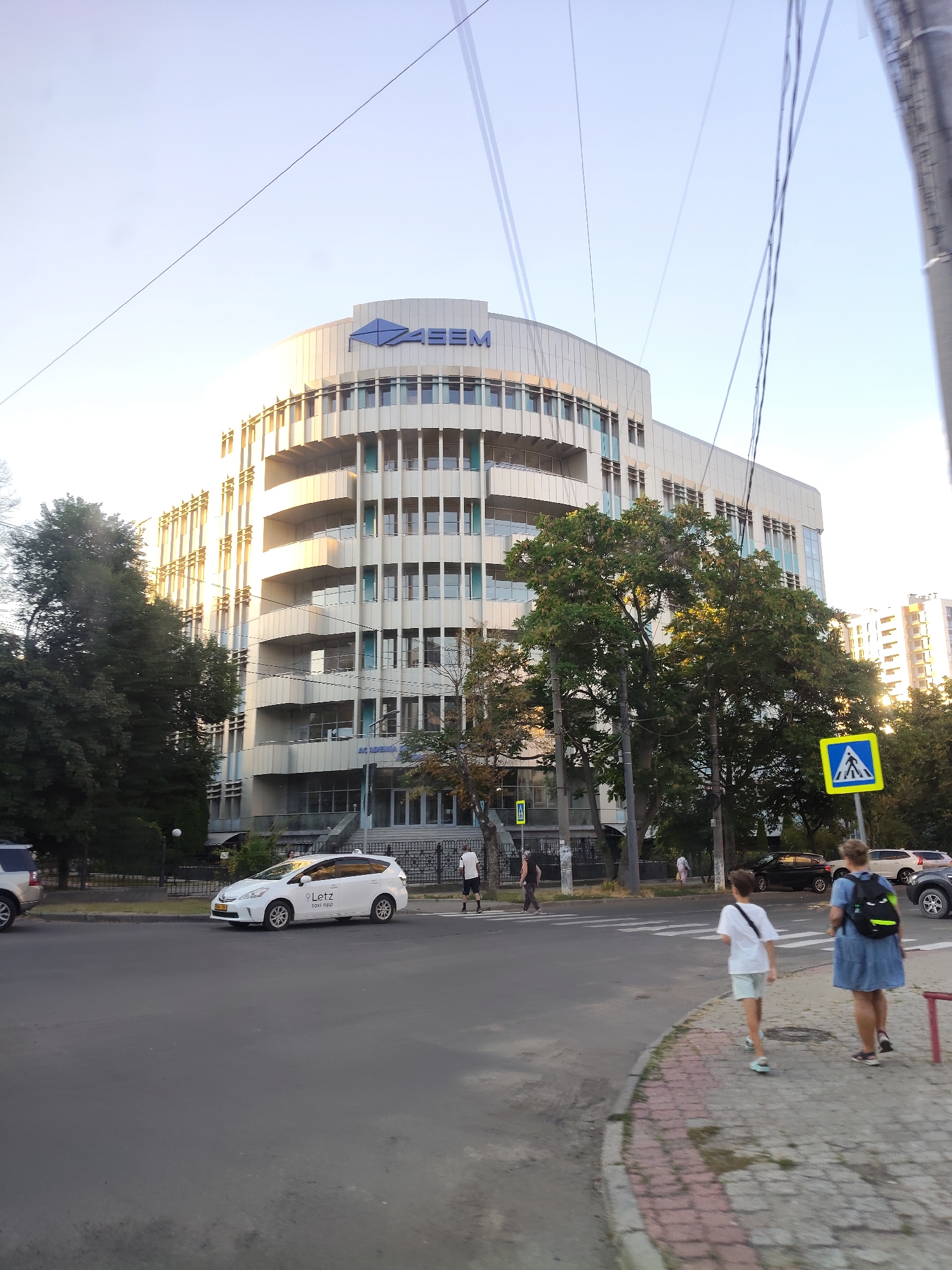 The image shows the ASEM (Academy of Economic Studies of Moldova) building in Chișinău, Moldova, viewed from a street. The building is a modern, multi-story structure with a curved facade, characterized by many windows and balconies. It is situated next to a road with traffic. A white Prius taxi with "Letz" branding is parked on the side of the road. Two pedestrians, a woman and a child, are walking on the sidewalk in the foreground, crossing a pedestrian crossing. A yellow pedestrian crossing sign is visible on a pole. Trees are present on the left and right sides of the image, and power lines crisscross the sky. The sky is clear and bright, indicating it is daytime.