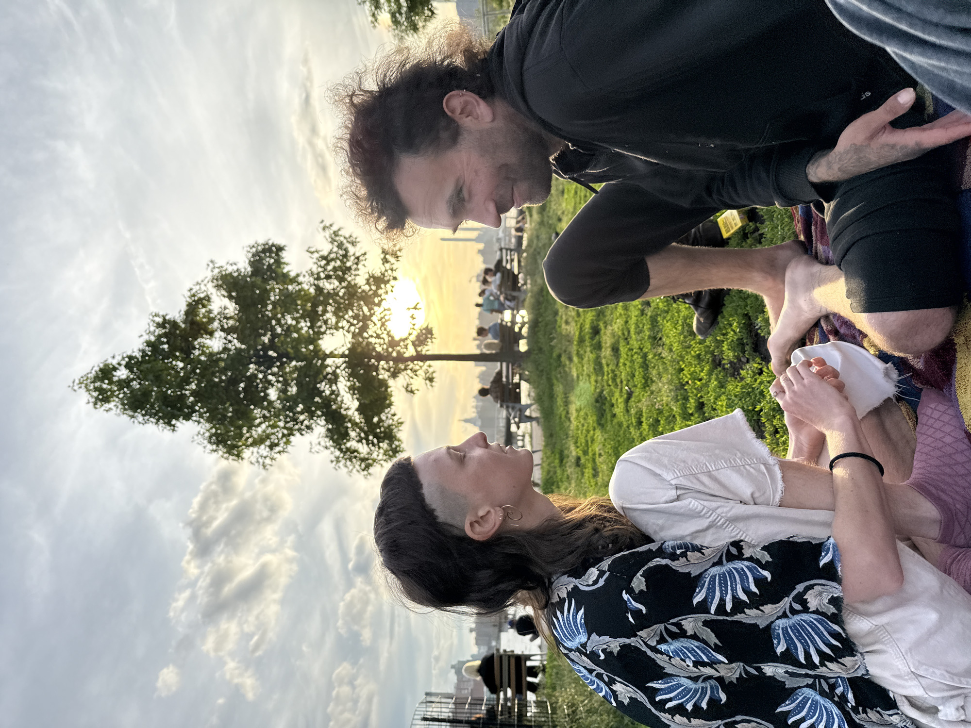 A man and a woman are interacting on a blanket in a park. The man, with curly brown hair, is kneeling and facing the woman, appearing to examine or touch her hands. He is wearing a black hoodie and dark pants, with bare feet. The woman, with short hair, is lying on her back, looking up at the man. She is wearing a patterned blouse, a white shirt, and light-colored pants. They are sitting on a colorful blanket laid out on the grass. In the background is a tree, a body of water, and a cityscape with people seated on benches. The location appears to be a park in New York City, United States of America, as indicated by the skyline visible in the distance. The scene is bathed in the warm light of sunset.