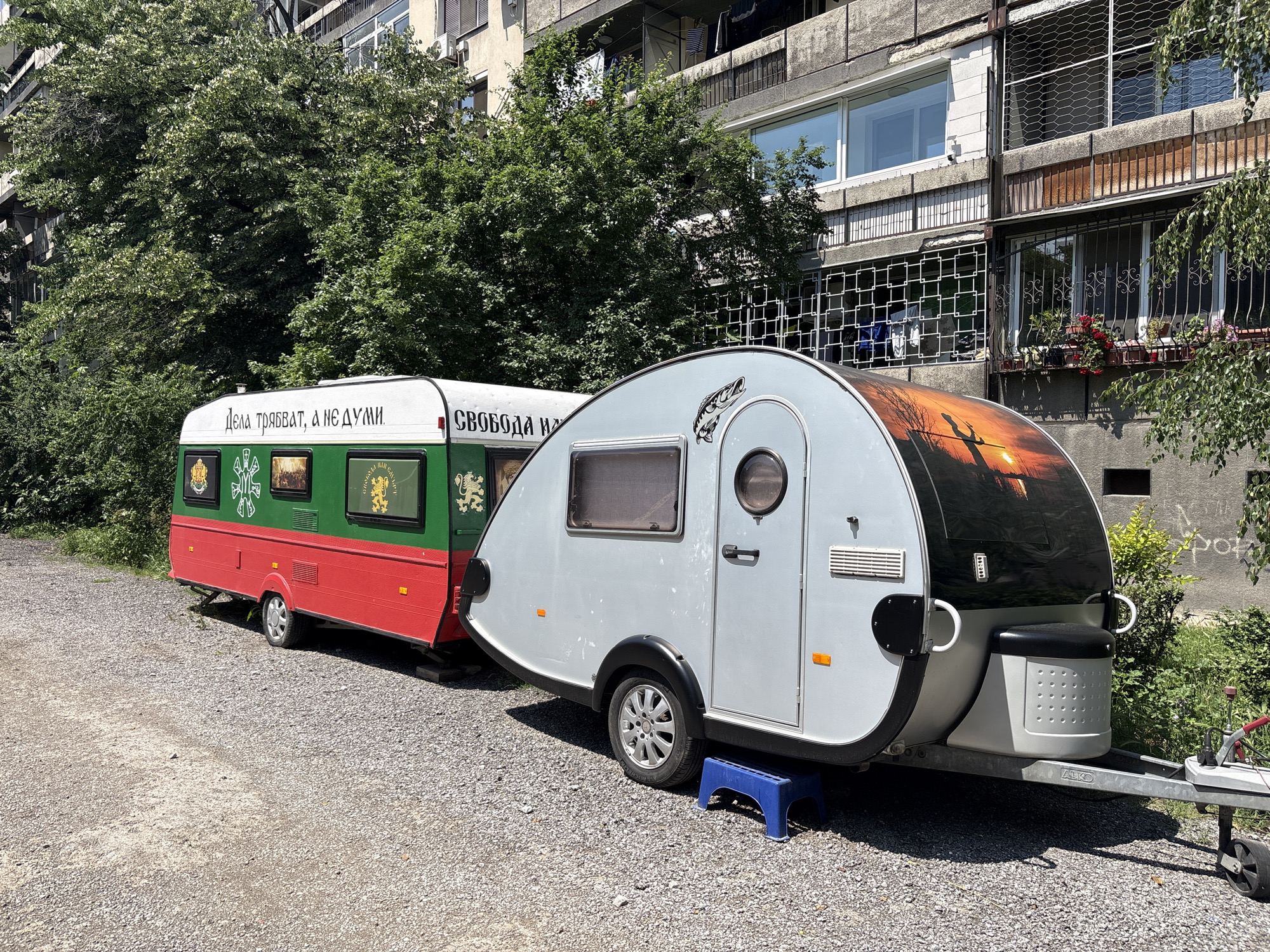 The image shows a two-part camper caravan parked on a gravel path in Plovdiv, Bulgaria. The leading camper is painted in silver with a sunset scene on its front. It has a fish design on its side. The towed camper is decorated in the colors of the Bulgarian flag (red, green, white) and features inscriptions and images. Behind the campers are apartment buildings, trees, and greenery. A blue plastic step stool supports the leading camper's wheel. The lighting suggests it's daytime with sunny conditions.