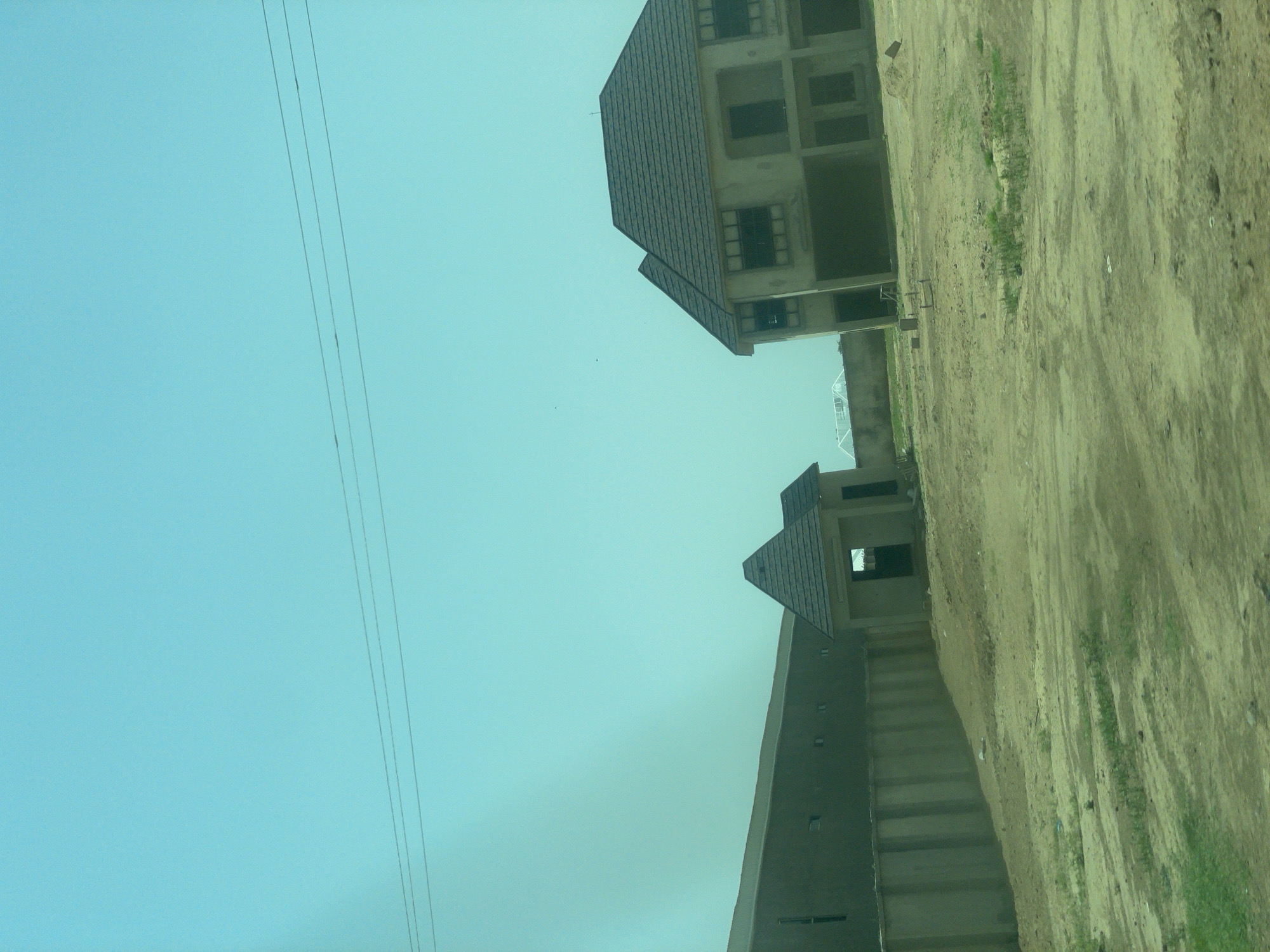 This is a photograph taken in Bauchi, Nigeria, showing a construction site with unfinished buildings. In the foreground, there is a sandy, uneven ground with patches of sparse green vegetation. A concrete wall runs horizontally across the mid-ground, separating the foreground from the structures behind it. To the left of the wall, a long, low building is partially visible, appearing to be a commercial or industrial structure with small, dark rectangular windows.
Further to the right, a smaller, single-story building with a pyramid-shaped roof stands. This building has a dark-colored roof and a dark opening that appears to be an entrance. Adjacent to this, and to its right, is a larger, two-story building that is also under construction. This building has a dark, tiled roof and a concrete facade. It features numerous large, dark rectangular window openings on both levels, indicating that the windows have not yet been installed. The building's overall design suggests it is a residential or commercial property.
In the background, the sky is a clear, pale blue, with three faint horizontal lines of what appear to be power or telephone wires stretching across the upper portion of the image. The scene depicts a developing area, likely within the city of Bauchi, characterized by ongoing construction projects. There are no people visible in the photograph.