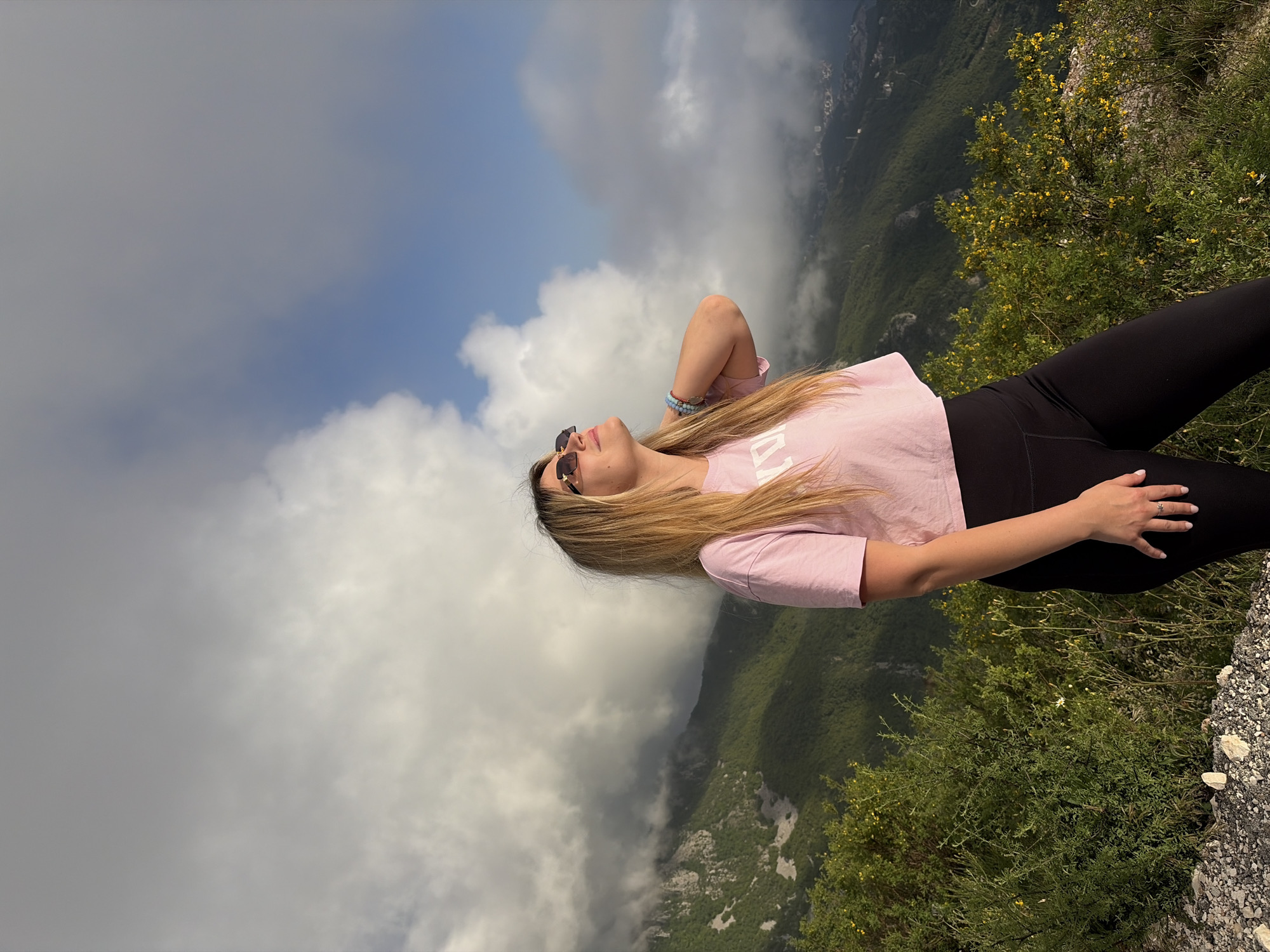 A woman is lying on a rocky edge overlooking a mountainous landscape, likely in Brajici, Montenegro, as indicated by the geotag. The sky is filled with puffy, white clouds interspersed with blue patches. The woman is wearing sunglasses, a light pink t-shirt, and black leggings. Her right arm is raised with her hand behind her head, and her left hand rests on her leg. The vegetation is thick, green shrubs and some small yellow flowers grow on the edge where she lies. In the distance, there are more green mountains partially obscured by clouds.