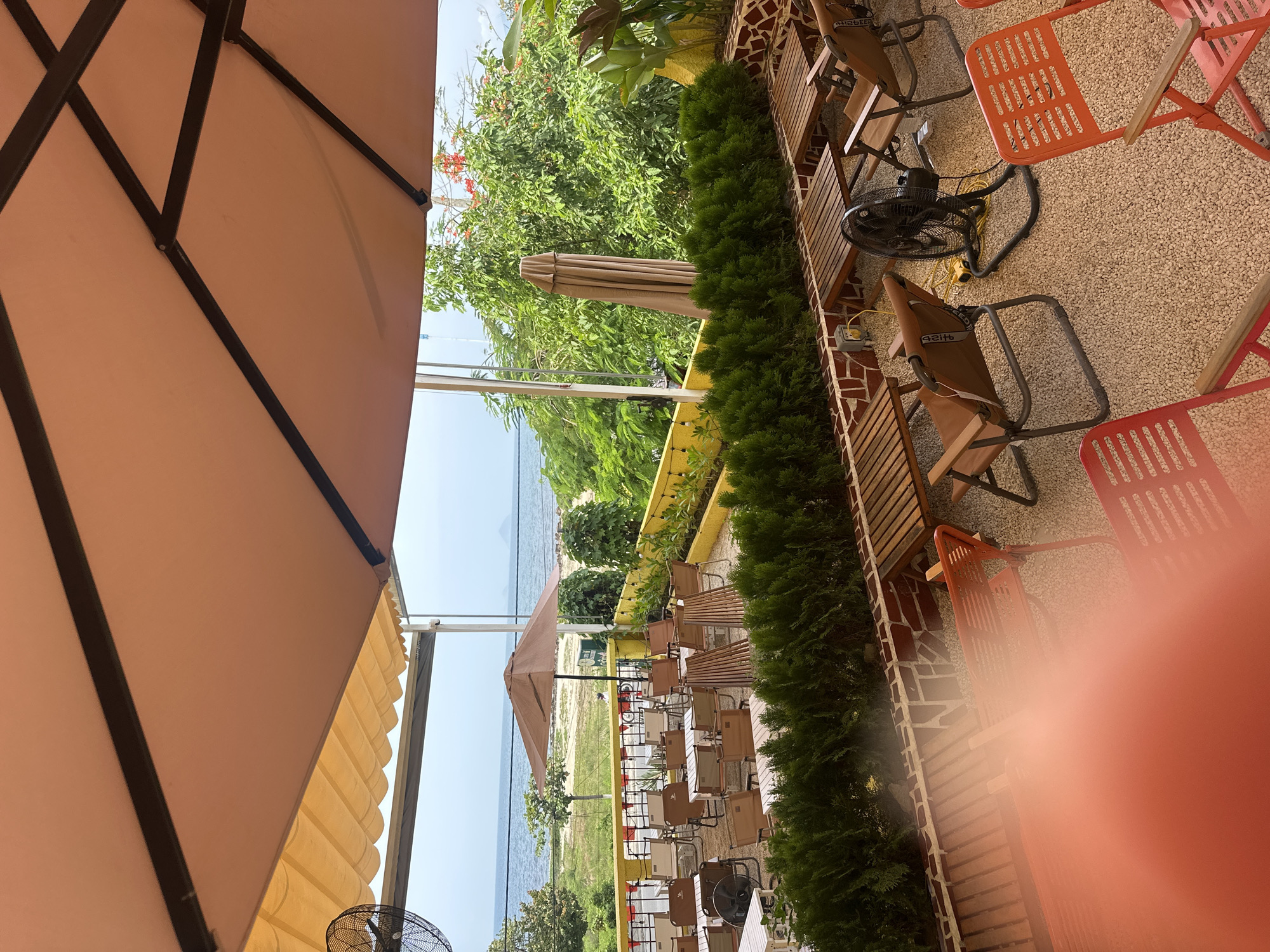 The image depicts an outdoor seating area of a cafe or restaurant located in Bac Nha Trang Ward, Vietnam, with a view of the ocean. Several tables and chairs are arranged on a gravel ground. In the foreground, there are orange metal chairs and tables with wooden tops. Further back, more tables and chairs, predominantly brown and beige, are set up. A large umbrella provides shade over some of these tables. Beyond the seating area, a low wall with yellow planters filled with green bushes separates it from a sandy area. In the distance, the ocean is visible with a faint horizon line. Lush green trees and foliage are present on the right side of the image, and a closed umbrella stands near a tall green plant. An industrial-style fan is positioned on the ground near some of the tables. The sky is overcast. The overall scene suggests a casual, open-air dining or relaxation space by the sea.
