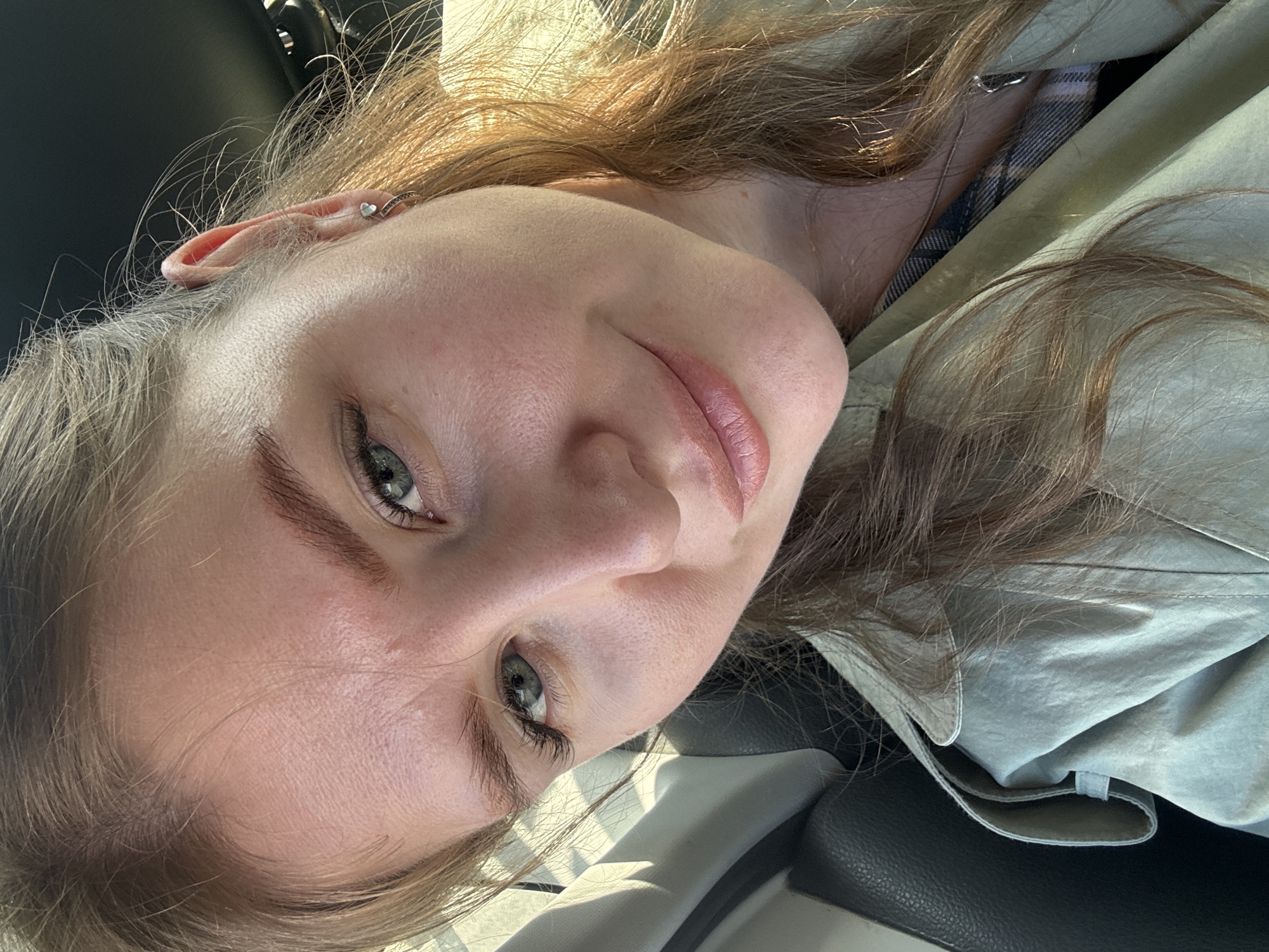 A close-up portrait of a woman's face. She has fair skin, light brown hair, and green eyes. She's wearing a light-colored garment with a collar. An earring is visible in her left ear. The background includes parts of a car interior. The location is likely Perm, Russia, based on the geotag.