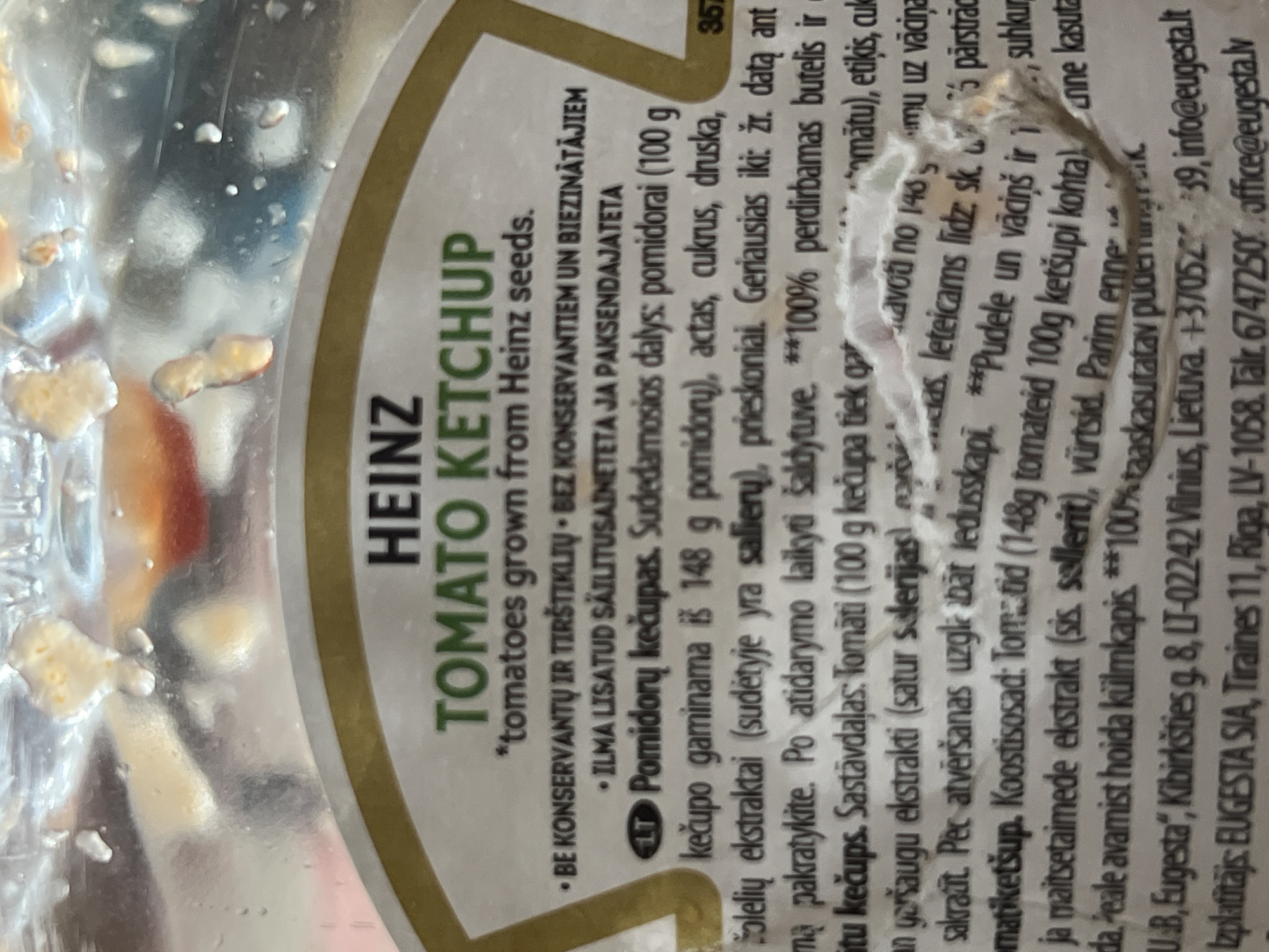 The image shows a label on a bottle of Heinz Tomato Ketchup. The label is white with black and green text. The text is in multiple languages, including English, Lithuanian, and Latvian. Some of the text is illegible due to damage to the label.
The image does not provide any information about the city of Daulėnai, Lithuania.