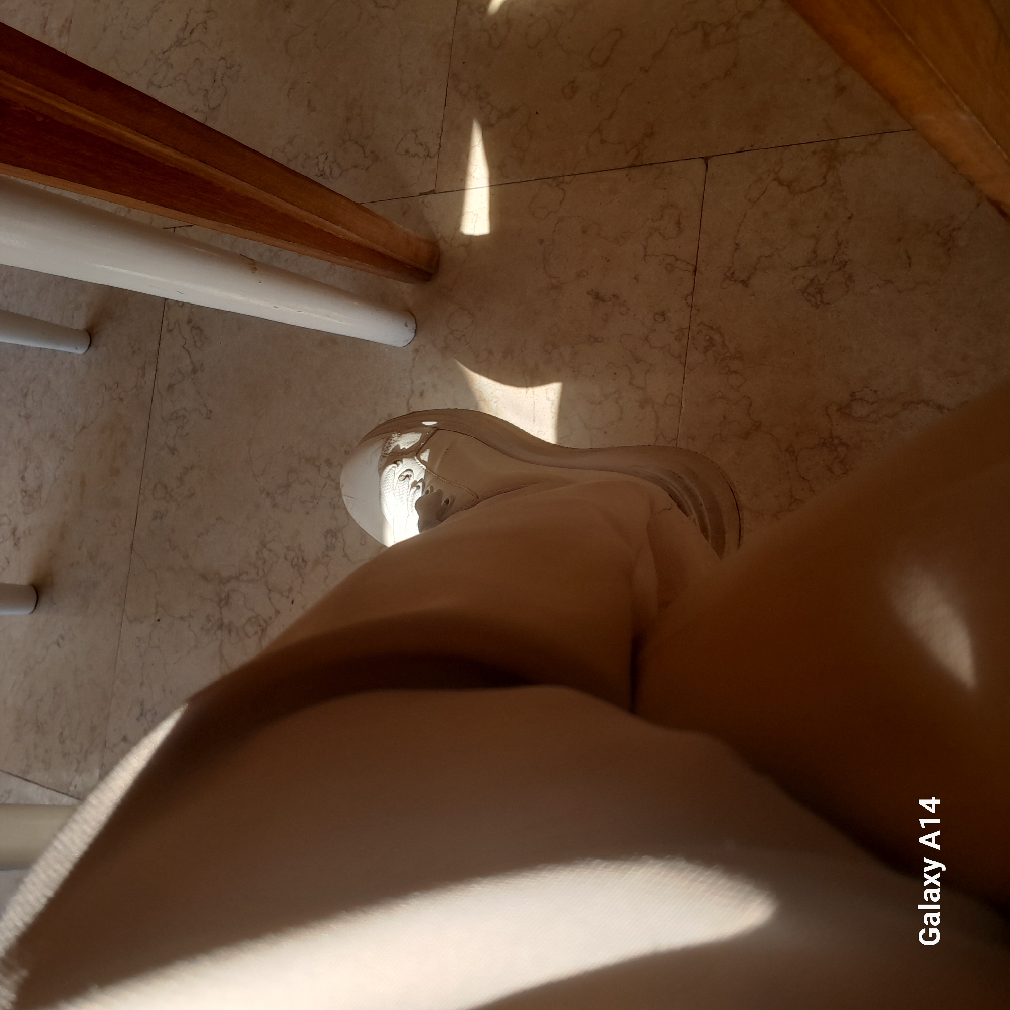 A person's leg, clad in light-colored pants and a beige sneaker, is visible from the knee down, positioned on a marble-tiled floor. Sunlight casts sharp shadows from unseen objects onto the floor and the person's leg, creating a dramatic play of light and dark. The background reveals the legs of two chairs, one white and one made of polished wood, suggesting an indoor setting, possibly a cafe or restaurant. The lower right corner of the image displays the text "Galaxy A14," indicating the device used to capture the photograph. The scene is devoid of any other discernible activity or interaction, focusing solely on the person's leg and the play of light and shadow on the floor.