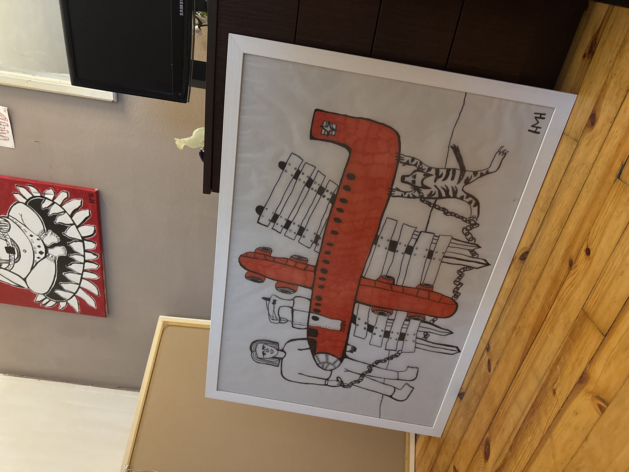 The media file captures an indoor scene in Sofia, Bulgaria, showcasing a collection of artworks and home furnishings. The primary focus is a large, framed drawing presented at an angle, resting on a wooden floor.
The central artwork features a graphic, black-and-white drawing on a light background, dominated by a large, stylized red object resembling a thick, elongated aircraft or submarine, detailed with black dots along its body. To the left, a simply outlined human figure is depicted, chained by the waist to the red object. To the right, a striped, tiger-like animal in black outline is also chained by its leg to the red object. Multiple abstract, rectangular structures, detailed with black circles and lines, surround the main red object, with some connected by chains, suggesting a mechanical or industrial theme. The drawing is signed with the initials "HWH" in the top right corner. The artwork is enclosed in a simple white frame.
To the left, a red canvas painting hangs on a light grey wall. This painting features a bold red background with a black-and-white stylized depiction, possibly of a face or mask with jagged teeth or spiky elements. The initials "RH" are visible in the bottom right corner of this canvas.
In the background, a dark brown wooden dresser or cabinet is visible, atop which sits a small, translucent white decorative object. A black flat-screen television with the "SAMSUNG" brand name partially visible is positioned above the dresser. The floor consists of warm-toned wooden planks.