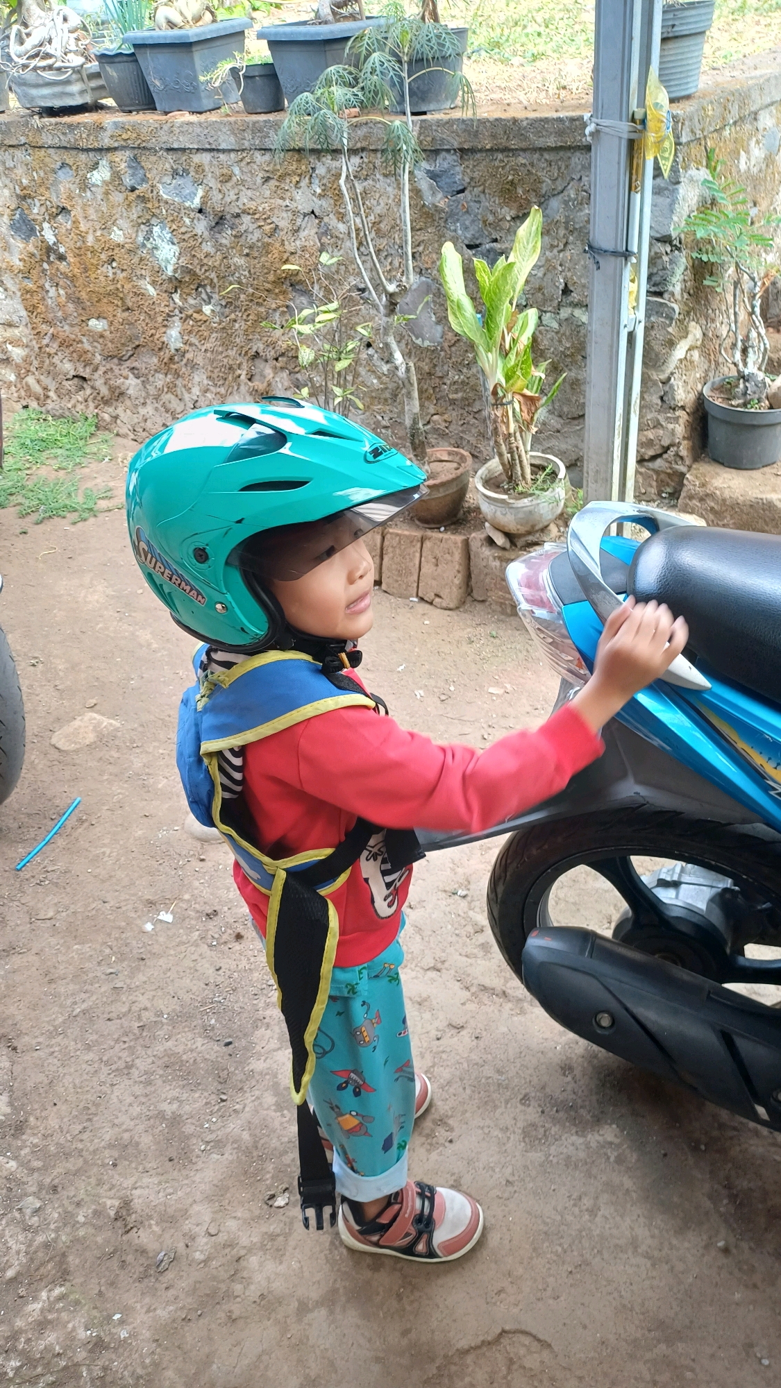 A young child, wearing a teal helmet with a "SUPERMAN" logo, a red long-sleeved shirt, and light blue pants with airplane and fish patterns, stands next to a blue and black motorcycle. The child is wearing pink and white shoes and a blue and yellow safety harness. The child's right hand is touching the motorcycle's seat. The scene appears to be outdoors on a dirt ground, with a stone wall and potted plants in the background. The location is identified as Wonokerso, Indonesia.