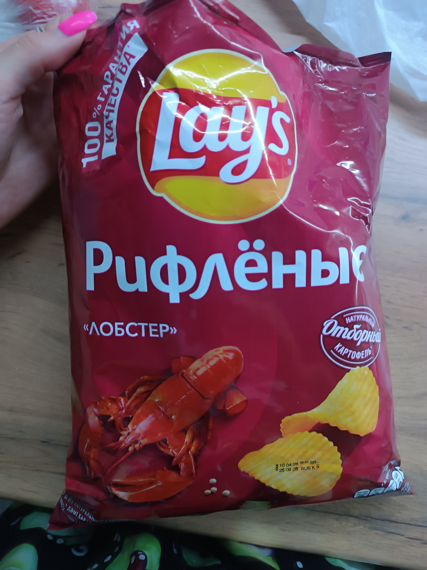A close-up shows a bag of Lay's brand "Rifljonje" (ridged) potato chips with "Lobster" flavor. The bag is predominantly red with the Lay's logo, the flavor name in Cyrillic, and an image of a lobster. The text "100% quality guarantee" is visible. There are images of potato chips as well. The chips are identified as "Naturally selected potato". A hand with pink nails is holding the bag. A production date is printed on the bag: 10 04 26 08:53 SR- 25 09 26 RUS K. The bag is on a wooden surface with a pattern of fruits or vegetables visible beneath.