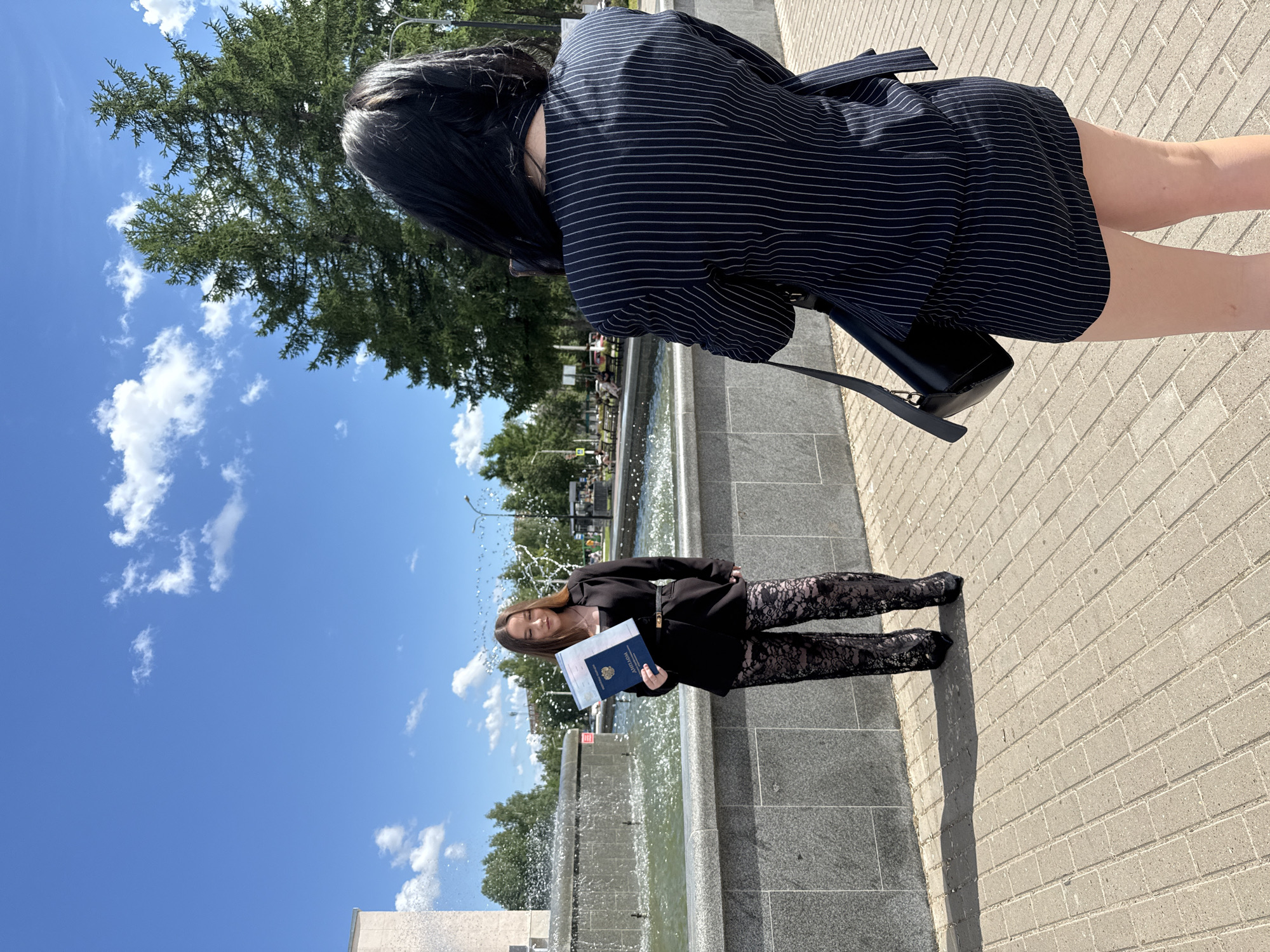In Chaikovsky, Russia, a young woman stands in front of a fountain, holding a diploma. She is wearing a black blazer, black lace-patterned pants, and black heels. The fountain has water spraying from it. Behind her, two evergreen trees are visible. Another woman, with dark hair, is positioned in the foreground, facing away from the camera and appearing to take a photo of the first woman. She is wearing a pinstripe suit and carrying a black purse. The scene takes place outdoors on a sunny day with a blue sky and some clouds.
