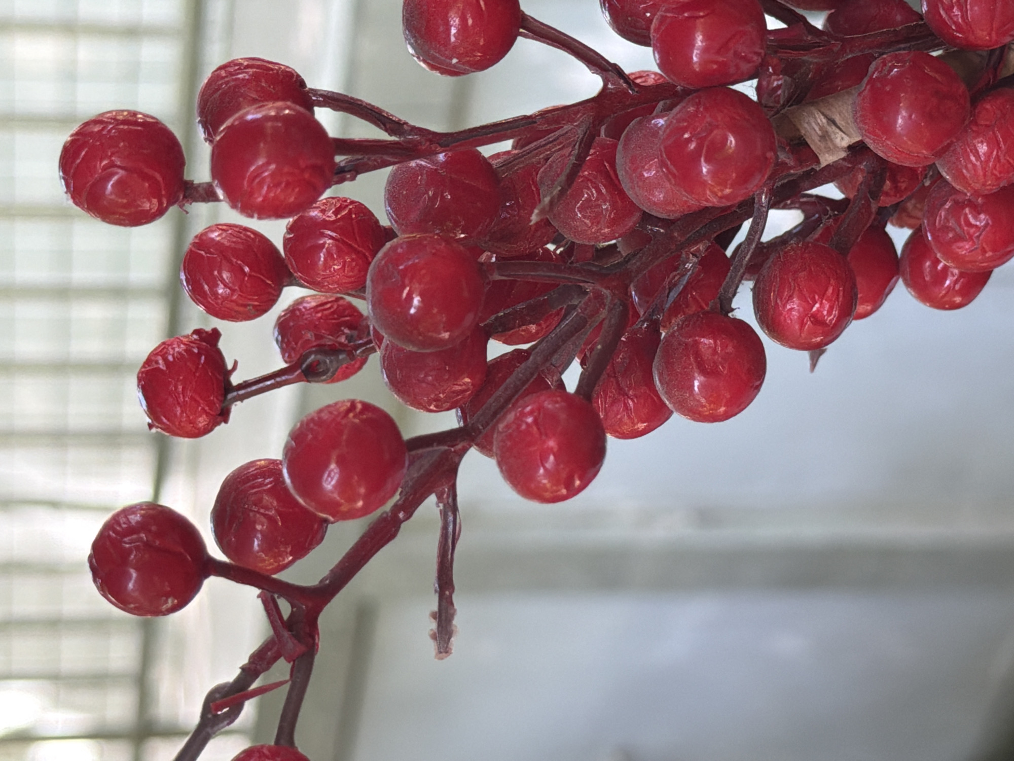 This is a close-up, vertically oriented, eye-level shot of a cluster of artificial red berries on dark red stems. The berries are round, with some showing slight indentations or wrinkles. The stems are thin and branched, with a matte finish. The background is a soft blur of pale gray and white, suggesting an indoor setting with diffused light, possibly a studio or a softly lit room. There are no people or specific identifiable landmarks from Kuta Selatan, Indonesia visible in the image.