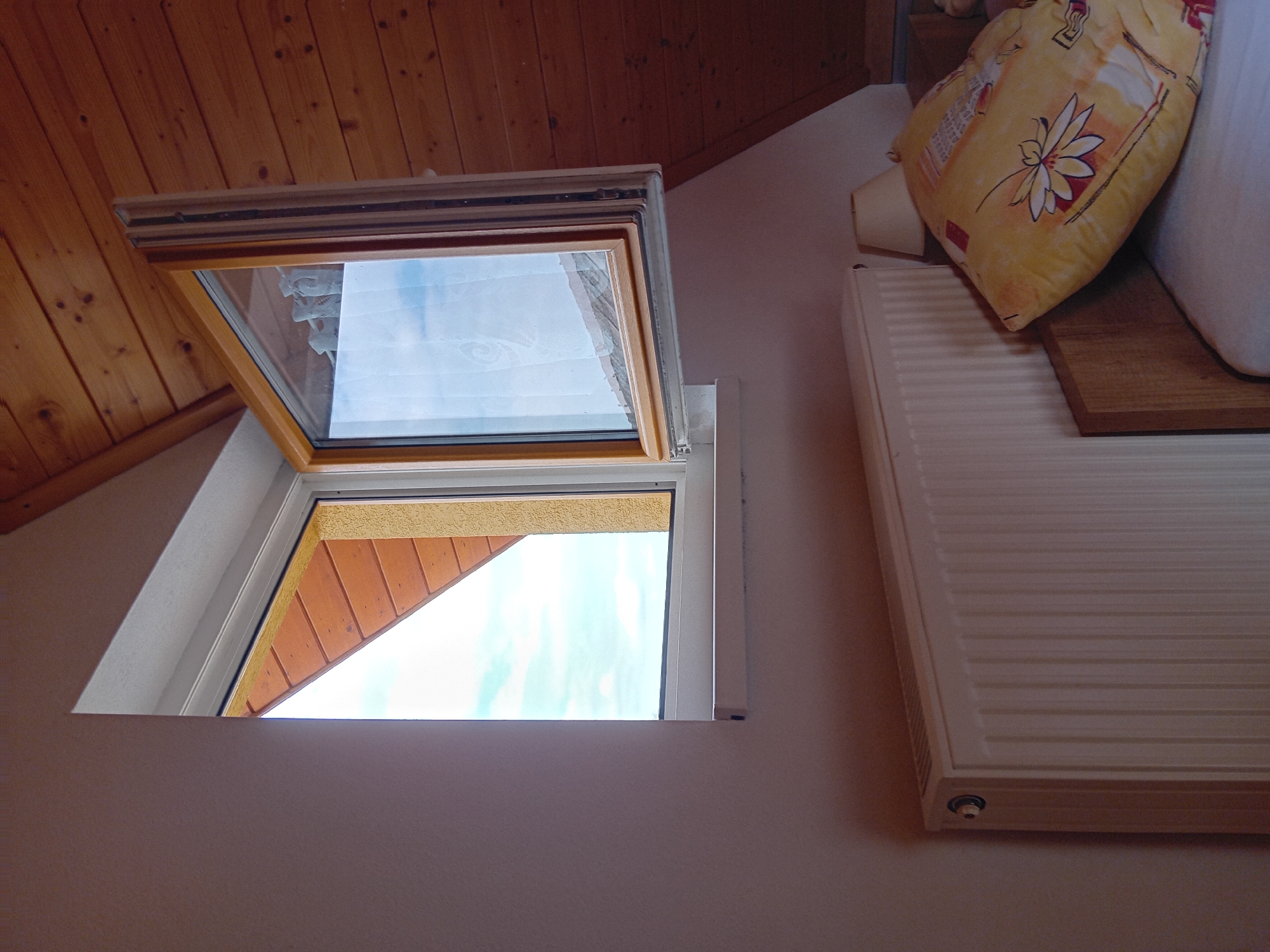 The image depicts an interior scene from a room in Zuberec, Slovakia. The view is from a low angle, focusing on a window and a radiator. The window is open, revealing a triangular section of the exterior with wooden roof eaves and a bright blue sky with some white clouds. The window frame appears to be made of wood with a light-colored trim. Adjacent to the window, the wall is painted a light, neutral color.
Below the window, a white radiator with vertical fins spans horizontally across the lower portion of the frame. To the right of the radiator, a wooden headboard of a bed is visible, and on the bed, a yellow pillow with a floral pattern is propped up. A lamp with a beige shade can be seen behind the pillow, on the right side of the image. The ceiling and upper walls are covered in warm-toned wooden planks, suggesting an attic room or a room with a sloped ceiling. The overall impression is one of a cozy, domestic interior.