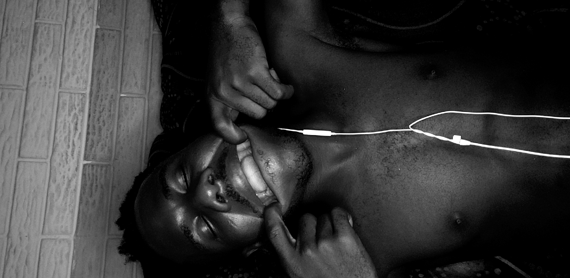 The image is a black and white photograph. A man is lying on his back. His face is upturned and he is holding his mouth open with his fingers. The man is shirtless, and an earphone cord is draped across his chest. The background is partially visible and appears to include a tile wall and some patterned fabric. The image is reported to be taken in Narayi, Nigeria.