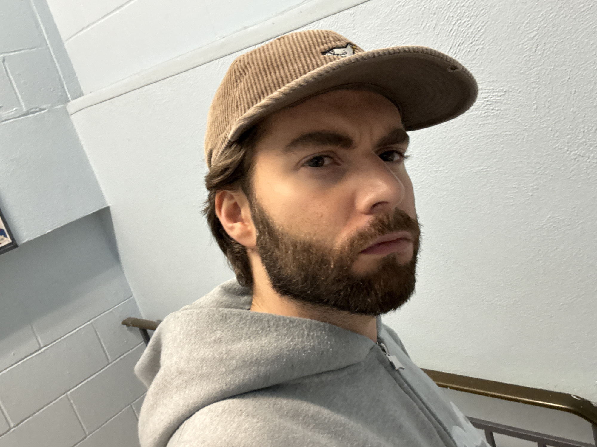 A medium, indoor shot depicts a bearded, white male wearing a tan corduroy cap and a gray hooded sweatshirt. The subject is positioned at an upward angle with a white wall with a slight texture behind him. To the left is a gray brick wall with a railing visible. No other individuals, objects, or actions are visible, and the location cannot be determined based on visual evidence.