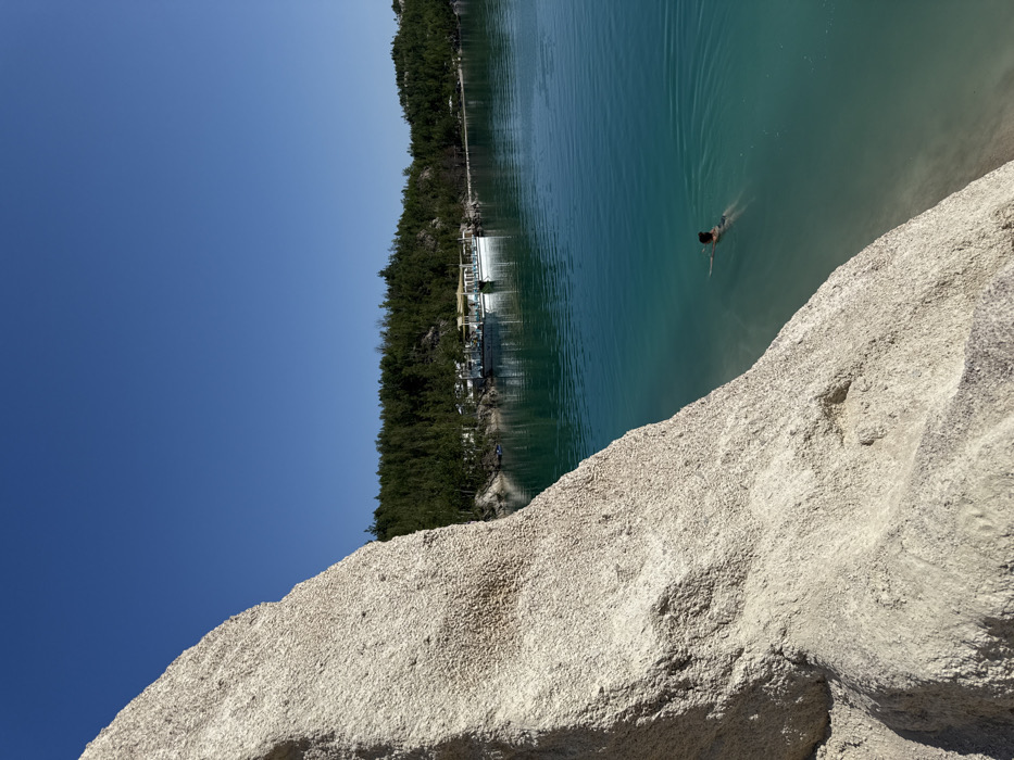 This image depicts a scene at a former quarry in Druzhba, Ukraine, specifically at the "Druzhba Quarry" or "Blue Lakes," characterized by its clear, turquoise water and white sandy/chalk banks.
The foreground on the left is dominated by a large, textured, light-colored (off-white/beige) rock or cliff face, angled upwards and to the right, showing coarse granular detail and natural contours. The majority of the right side of the frame is occupied by a large body of water, exhibiting a distinct turquoise to emerald blue hue, appearing calm and reflecting the sky and distant land.
In the mid-ground, a single individual is observed swimming in the water, with their head and upper torso visible above the surface, oriented away from the camera. In the background, across the water, a dense line of dark green trees (likely coniferous) defines the distant shoreline. Along this tree-lined bank, several light-colored structures are discernible, including two white, pontoon-style boats moored near the shore; one boat features a light-colored canopy. The sky above is clear and uniformly bright blue, indicative of a sunny day.