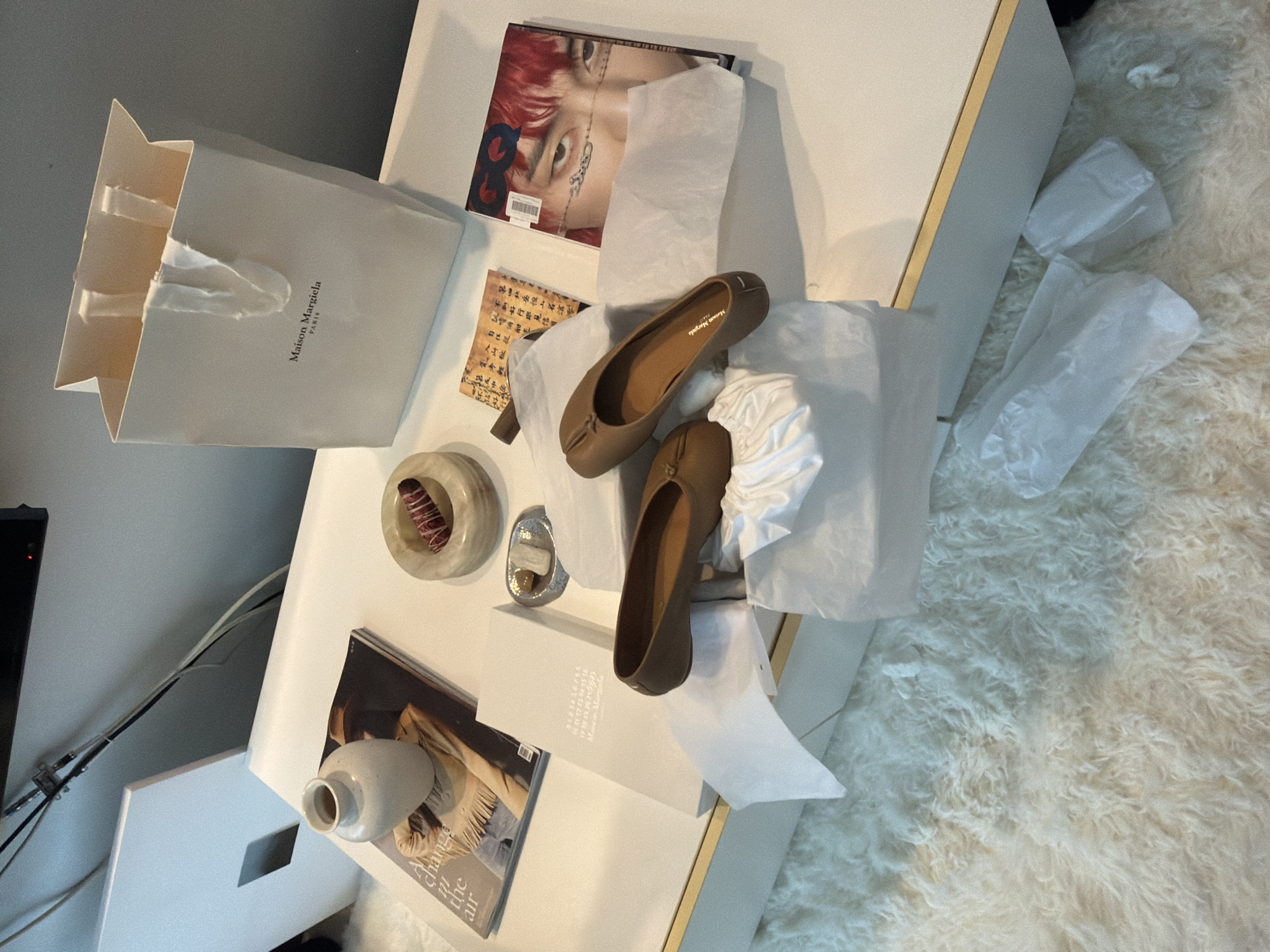 This media file captures an indoor still-life scene, likely in Seoul, South Korea, featuring fashion items and decorative elements arranged on a white table next to a fluffy white rug. The primary subjects are two light brown Maison Margiela Tabi ballet flats, distinctive for their split-toe design, placed on white tissue paper. One shoe is fully visible, displaying "Maison Margiela" printed on its insole, while the other is partially obscured.
Adjacent to the shoes is a white Maison Margiela shopping bag with "Maison Margiela PARIS" printed on its side. Also on the table are several books or magazines; one features a close-up of a person with red hair and face tattoos on its cover, and another has text that reads "Change the air" along with an image of a person. A smaller book or card displays "Maison Margiela" and a sequence of numbers (1 to 22).
Decorative objects on the table include a small white ceramic vase, a circular light-colored stone dish containing a reddish, cylindrical item (possibly sage or incense), and a small silver dish. The background features a plain grey wall with some visible wires on the left side, suggesting a functional or utility area in the room. The overall impression is one of a curated display, possibly showcasing new purchases or styled decor. The lighting is even, suggesting an indoor setting without strong natural light sources.