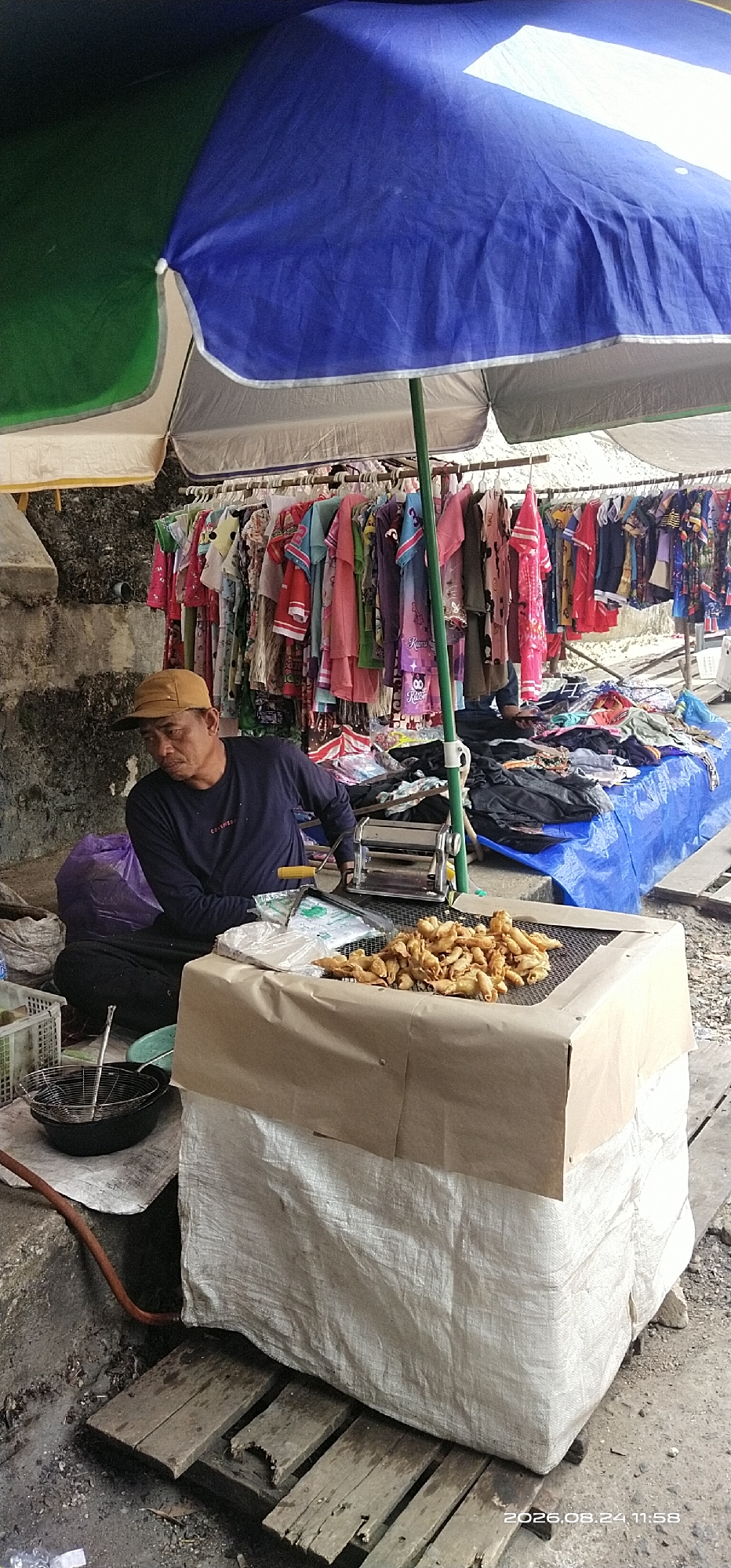 A man sits cross-legged under a large blue and green umbrella at an outdoor market stall in Tamiang Layang, Indonesia. He is wearing a dark long-sleeved shirt and a tan baseball cap. In front of him is a metal pasta maker and a grill covered with fried food items, possibly bananas. Behind him, clothing is displayed on a rack for sale. The stall is set up on wooden planks, with a blue tarp covering some of the goods. The background shows a rough stone wall and some bags. A frying pan with a wire basket is visible to the left. The overall scene depicts a vendor operating a street food stall.
