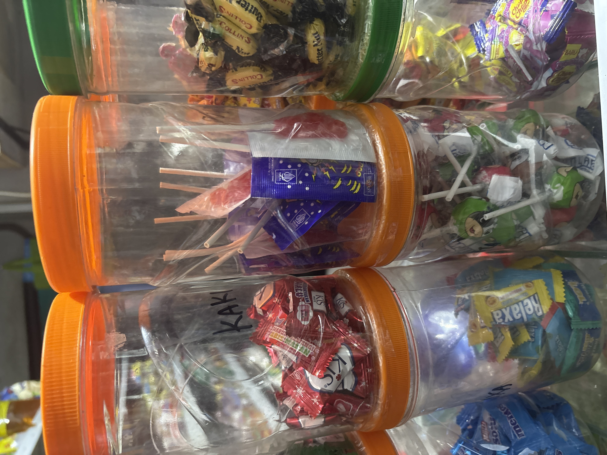 The image shows a close-up view of several clear plastic jars filled with various candies and lollipops. The jars have colorful lids, with orange and green being prominent. Some of the candies are individually wrapped in colorful packaging. In one jar, "KAKI" is written in black marker on the inside. The lighting suggests an indoor setting, possibly a shop or market stall. The presence of Indonesian candy brands like "Relaxa" and "Kis" suggests the location is likely North Jakarta, Indonesia, a common place for such retail displays. The scene is static, with no people or active interactions depicted. The focus is solely on the collection of confectionery items.