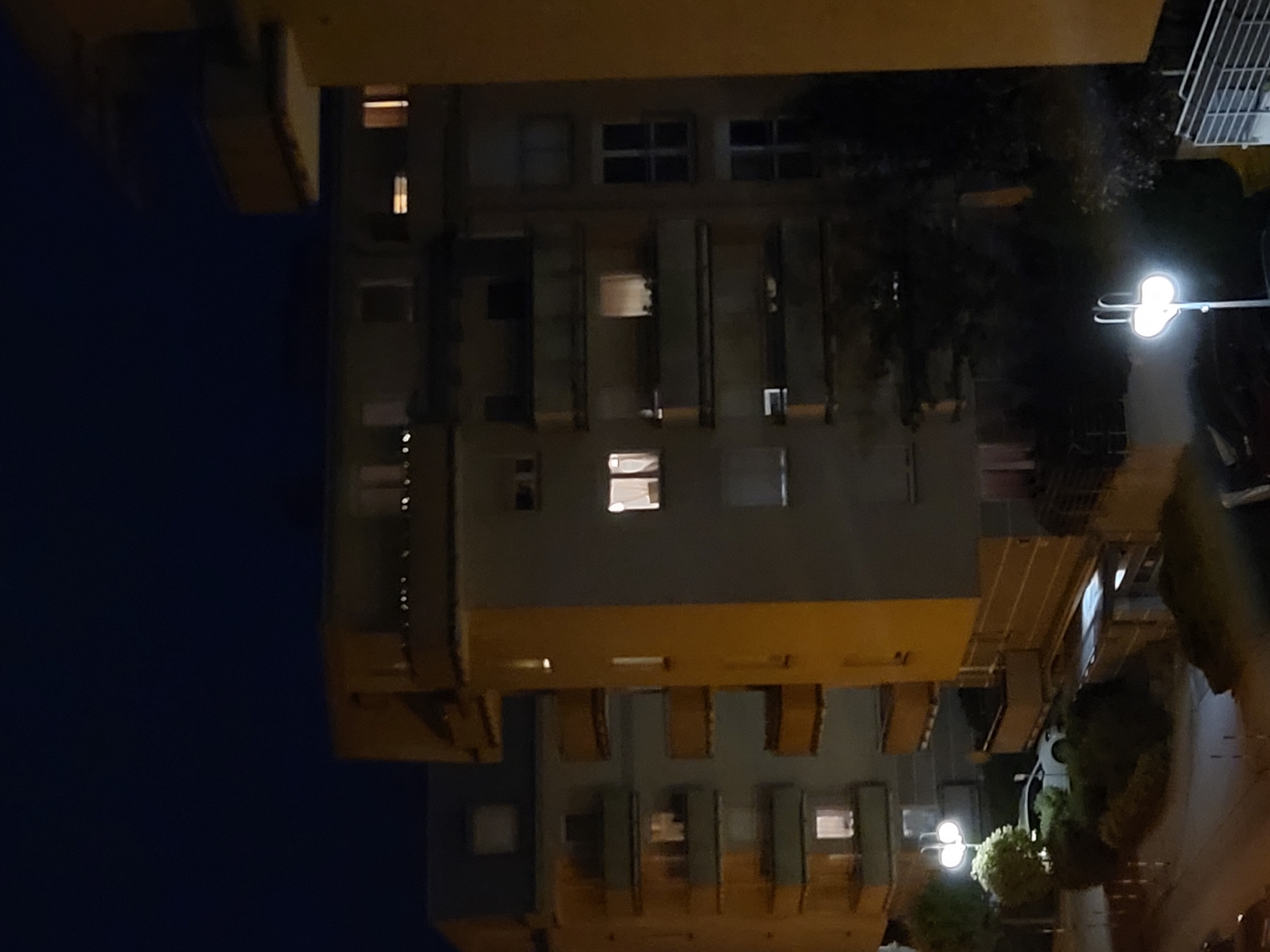 The image depicts an exterior view of apartment buildings at night in an urban setting. The buildings are illuminated by lights from windows and streetlights. In the foreground, a streetlight with two globes illuminates a small white car parked amidst green foliage. The architecture of the buildings is characterized by balconies with railings. The sky is dark, indicating nighttime.