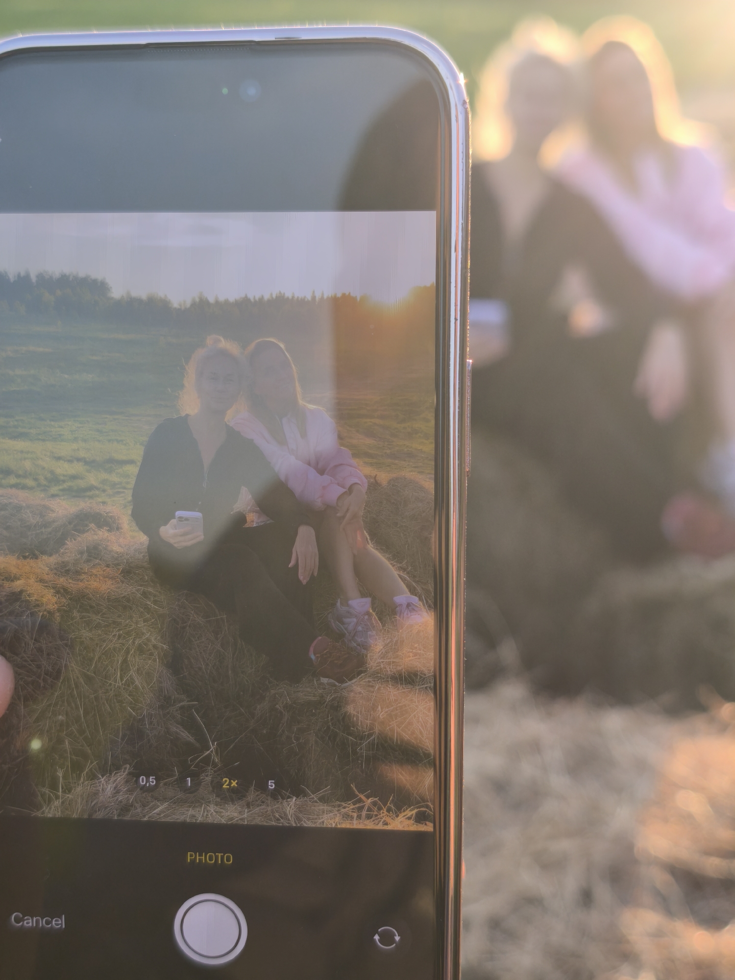 The image captures a "photo within a photo" scenario, where a smartphone is held up to photograph a scene, with its screen displaying the live camera feed. The primary focus is on the smartphone, likely an iPhone given its distinct notch/Dynamic Island design, held vertically by a hand partially visible in the bottom left corner.
On the phone's screen, two young women are depicted sitting closely on hay bales in an open field. The woman on the left, with blonde hair, wears a dark zip-up top and holds a white smartphone, looking directly at the camera with a serene expression. Her companion on the right, wearing a light-colored (possibly pink) jacket, has her arm around the first woman's shoulder and also looks at the camera with a slight smile. The background on the screen reveals a vast field with scattered hay bales, lush green grass, and a distant line of trees under a bright, warm sun, creating a significant lens flare and a soft, golden glow across the image.
Behind the phone, the actual scene being photographed is visible but intentionally blurred, emphasizing the phone's screen. The shapes of the two women, the hay bales, and the intense golden light are discernible, mirroring the content displayed on the screen.
The time of day is clearly the golden hour, either sunrise or sunset, indicated by the intense warm light and the sun's low position, which creates the prominent lens flare. The setting is an outdoor, rural landscape, specifically located in Стеклянный, Russia, characterized by agricultural fields and natural elements. The activity is the act of taking a photograph, capturing a moment of friendship and relaxation in nature.
Visible text on the phone's camera interface includes zoom options "0.5", "1", "2x", "5" (with "2x" highlighted), the word "PHOTO" indicating the mode, a circular shutter button, "Cancel" option, and a camera-switch icon.