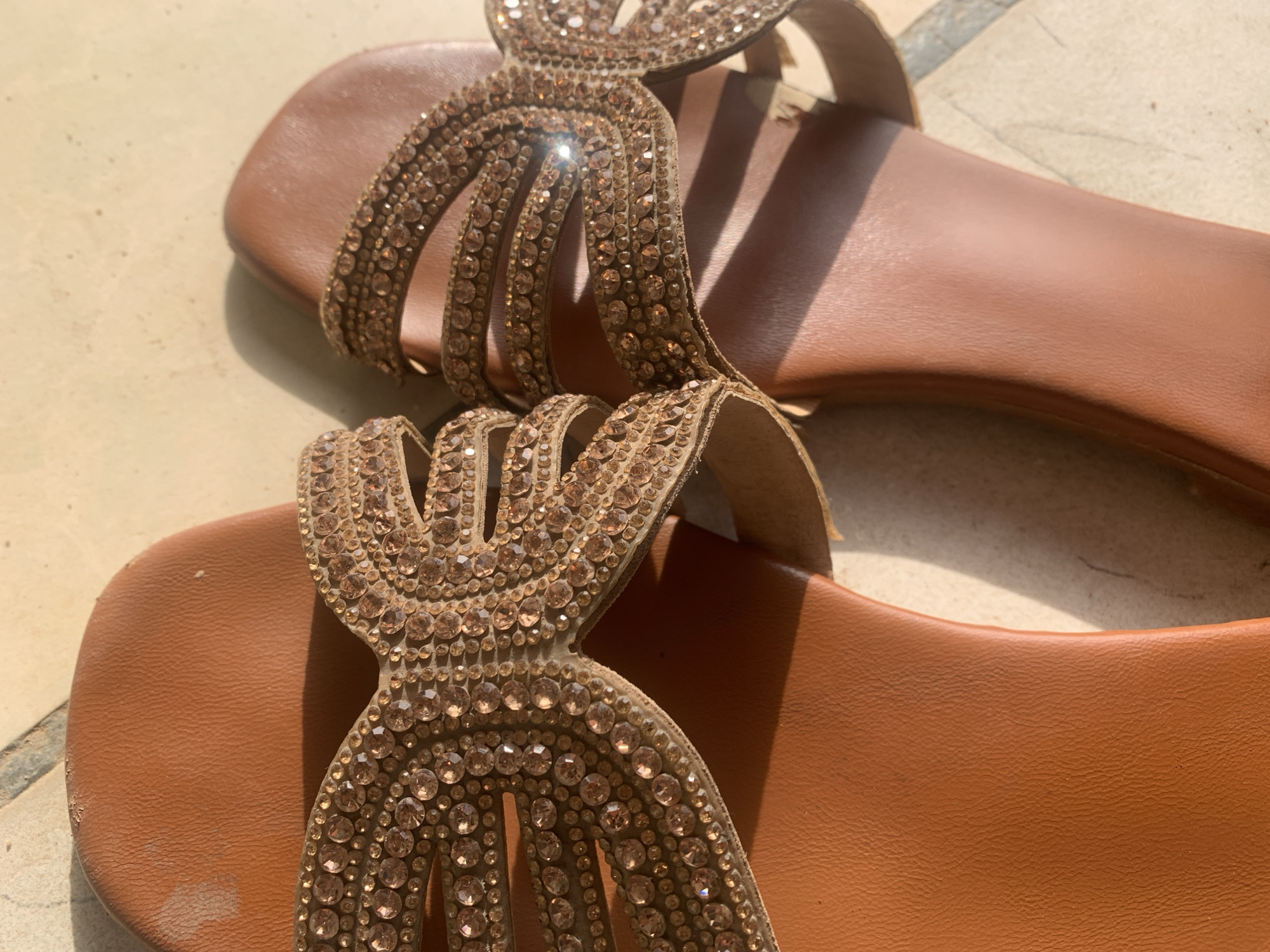 The image shows a pair of brown, jeweled sandals sitting on a light-colored tile surface. The sandals are adorned with numerous small, amber-colored rhinestones or beads. One sandal is positioned closer to the viewer, displaying the upper part with the decorative embellishments. The second sandal is situated behind and slightly to the right, partially obscured but still showing the similar bejeweled pattern. The setting suggests an indoor or outdoor tiled area. The lighting indicates it is daytime, with strong sunlight casting shadows. There is no evidence to indicate the specific location in the city of New Stadium, Nigeria. No people, interactions, or actions are visible.