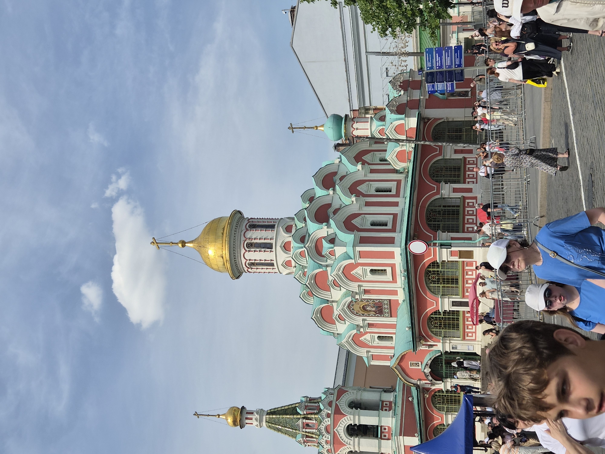 The image captures the Kazan Cathedral in Moscow, Russia, under a bright blue sky with scattered white clouds. The cathedral, centrally positioned, exhibits a red and white neoclassical facade with arched windows, teal-green roofs, and multiple domes. The largest central dome is a golden onion dome topped with a cross. To its left, a smaller bell tower is crowned with a similar golden onion dome and cross. A smaller teal dome with a cross is visible to the right. A religious mosaic icon adorns the facade below the main dome.
In the foreground, a crowd of people occupies the lower half of the frame. On the far left, a young person with brown hair is partially visible, facing right. To their right, an older woman wearing a white baseball cap and sunglasses, dressed in a blue t-shirt, looks upward and right. A younger woman beside her, also in a white cap, sunglasses, and a blue t-shirt, faces forward.
Metal crowd control barriers stretch across the middle ground, delineating pedestrian areas. Numerous individuals are seen walking or standing behind these barriers; some appear to be entering or exiting the cathedral's main entrance. A blue canopy tent is on the far left, and a red umbrella is present further back in the crowd. A blue informational signpost with white text and a red circular regulatory sign are located to the right of the cathedral's entrance. Overhead string lights or decorative elements are suspended above the street on the right side. Green foliage from a tree is visible at the far right edge. The ground surface is paved, featuring white markings, possibly cobblestone. A plain white building stands behind the right section of the cathedral, and a pink building is partially visible behind the left bell tower. The scene is illuminated by daylight.