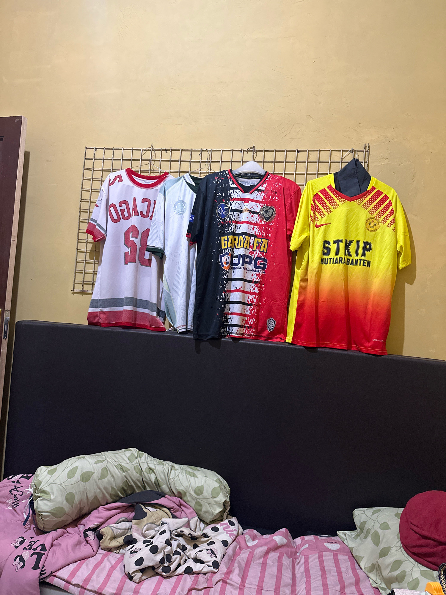The image shows four t-shirts hanging on a metal rack against a yellow wall. From left to right, the t-shirts are: a white t-shirt with red numbers and trim, a light blue t-shirt with white markings, a black and red t-shirt with various logos and text, and a yellow and red gradient t-shirt with text. Below the t-shirts is a dark gray upholstered headboard or backrest of a bed. On the bed in the foreground, there are several pillows and blankets, including a long bolster pillow with a green leaf pattern, pink striped sheets, and a pink blanket with black and white polka dots and images of Minnie Mouse. A red hat is also visible on the right side of the bed. The image appears to have been taken indoors. The location is identified as Saruni, Indonesia, although no specific landmarks within Saruni are visible in the image.