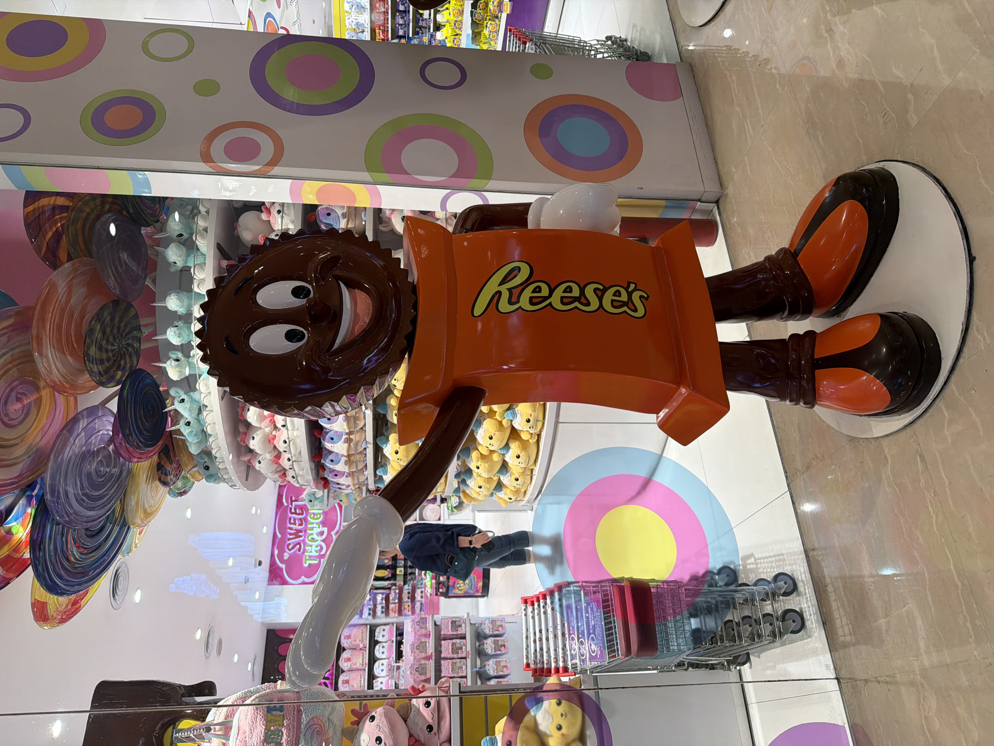 The photograph shows a large, cartoonish Reese's Peanut Butter Cup statue in what appears to be a candy store. The statue has a smiling face, an orange wrapper with the "Reese's" logo, and brown legs ending in oversized shoes. The statue is standing on a white base.
The store's interior features colorful decorations including large candy shapes and displays of plush toys and candy merchandise. There are shopping carts present in the store. In the background, a person wearing a blue shirt and jeans is looking at a phone.
Based on the presence of the "Sweet Thooth" sign, the store is most likely Sweet Thooth located in the Beachwalk Shopping Center in Kuta, Indonesia.