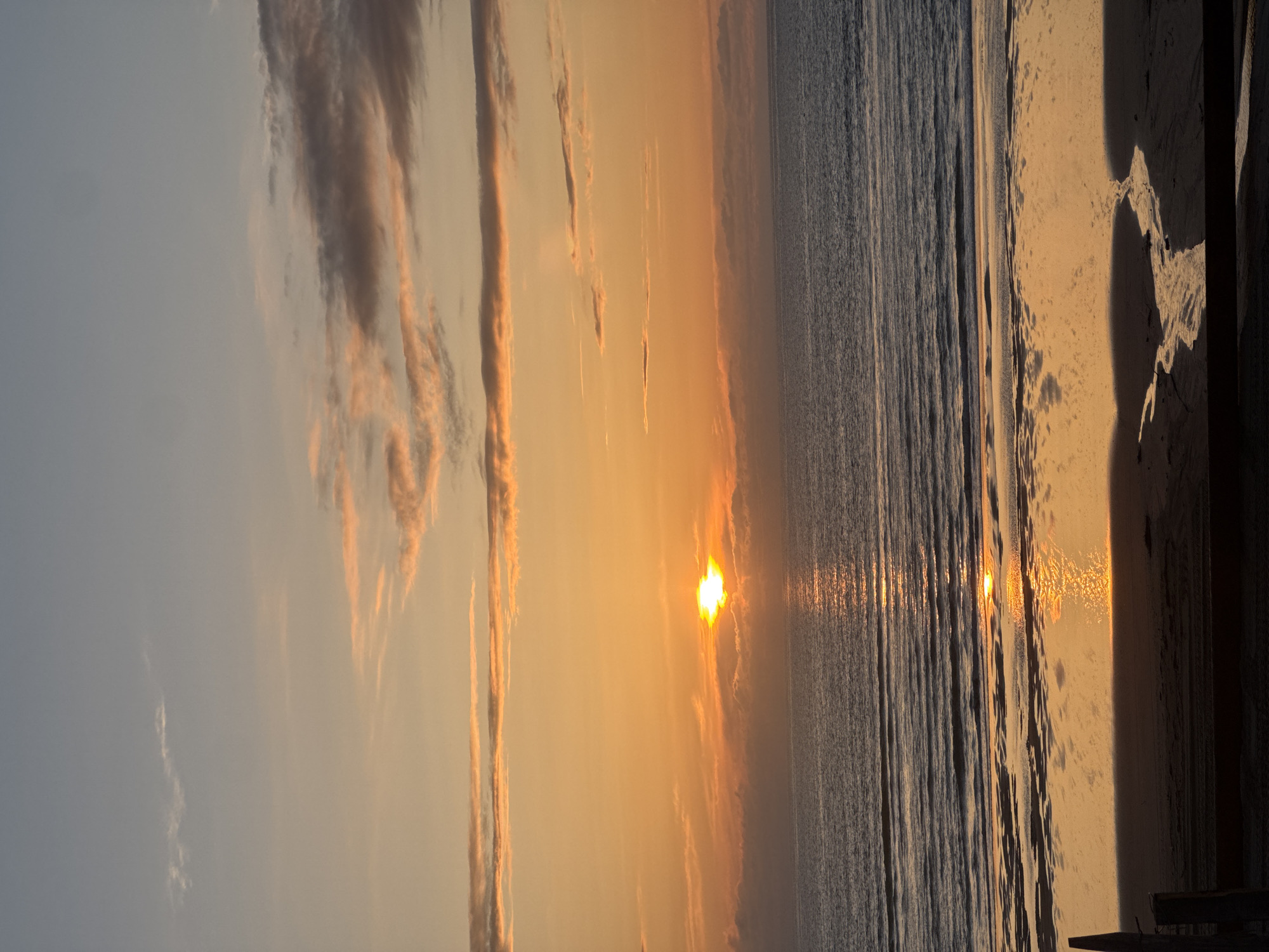 The image depicts a sunset over the ocean in Soulac-sur-Mer, France. The sun is low on the horizon, partially obscured by clouds, casting an orange glow across the sky and water. The beach is in the foreground, with some standing wooden palings visible on the right side of the image. The ocean has small waves. The sky has a mix of grey and golden clouds.