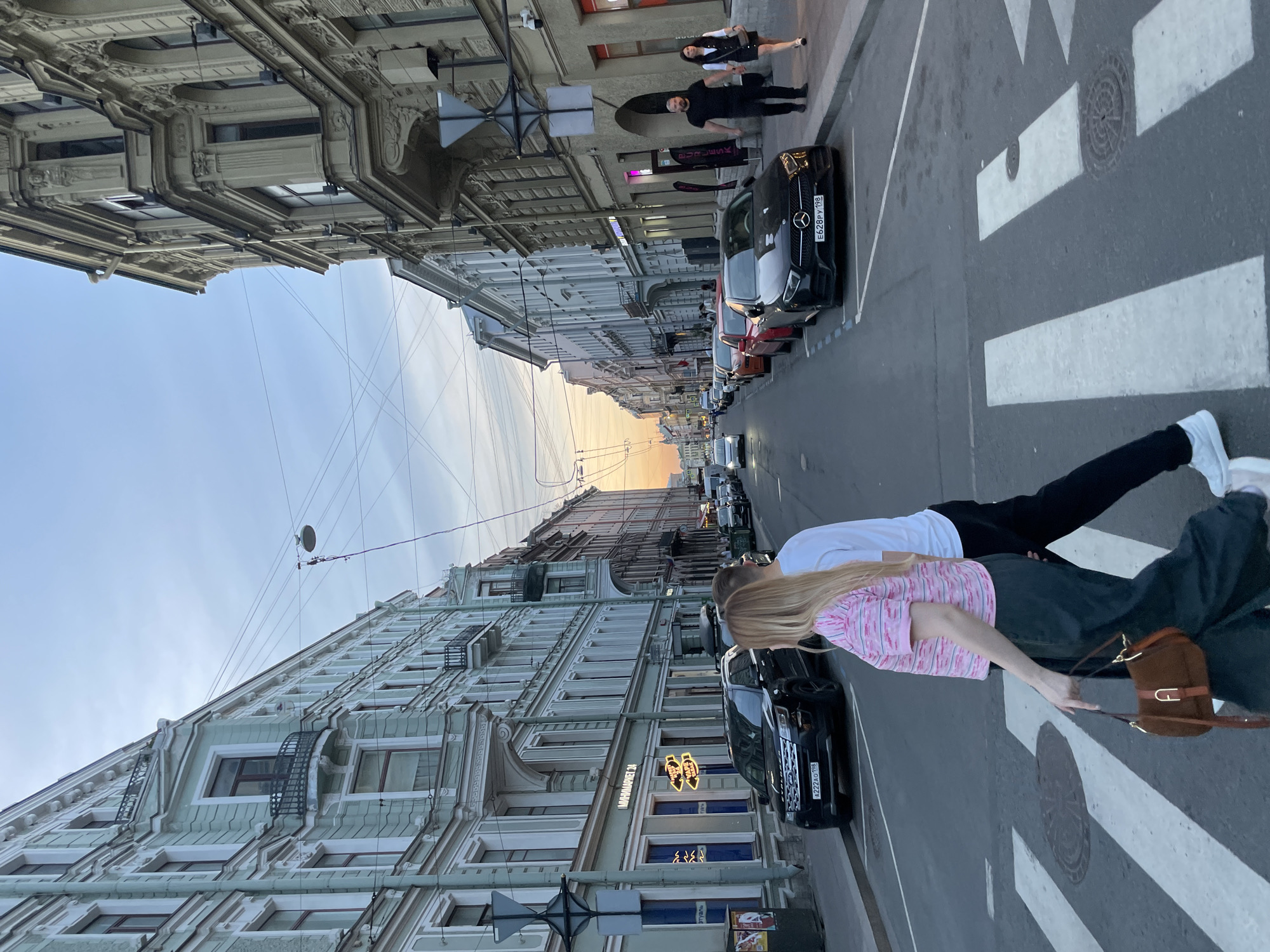 This image captures a street scene in Saint Petersburg, Russia, at dusk, characterized by the gradient of the sky from light blue overhead to orange and pink on the horizon. The presence of Russian license plates with the "198" region code confirms the location.
In the foreground, a white pedestrian crosswalk spans the asphalt street. Two individuals are traversing the crosswalk from left to right. The person on the left is a woman with long blonde hair, wearing a pink and white striped short-sleeved top and dark wide-leg trousers. She carries a brown shoulder bag with a gold "A" shaped clasp. Beside her, on the right, is a man with short dark hair, wearing a white t-shirt, dark trousers, and white sneakers.
The street is lined on both sides by multi-story, historic European-style buildings. On the left, the buildings are predominantly light teal with white trim, featuring ornate balconies and cornices. The ground floor of one such building displays the illuminated sign "МИНИМАРКЕТ 24" and yellow arrow signs. A black Ford Explorer SUV (license plate A222AO 198) is parked along the curb on the left, facing away from the viewer, with a cargo box on its roof. A dark grey utility pole with various signs stands nearby.
On the right side of the street, the buildings are lighter in color, possibly beige or light grey, also featuring ornate details and arched ground-floor windows. One shop sign is partially visible, reading "PUR LESK." A black Mercedes-Benz CLS (license plate E628PY 198) is parked facing the camera on this side. Further down the sidewalk on the right, another couple is walking towards the viewer: a bald, bearded man in a black t-shirt and trousers, walking alongside a woman with dark hair wearing a white shirt, black sleeveless top/dress, and black skirt.
Numerous overhead wires crisscross the sky, indicative of urban infrastructure. A streetlamp with a circular white shade hangs from these wires on the left side of the frame. The street recedes into the distance, with a line of parked and moving vehicles, and the sky's warm hues casting a soft light over the urban environment.