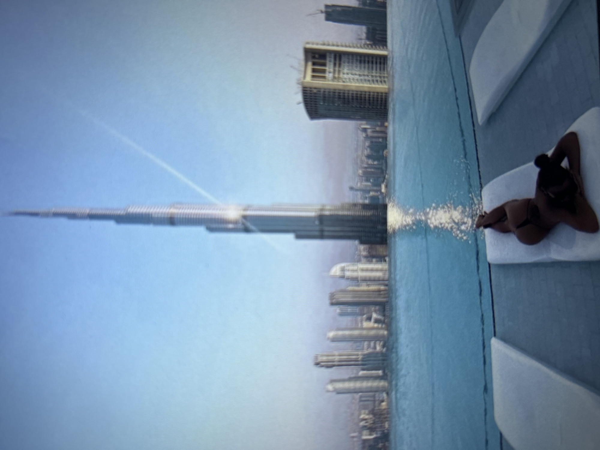 The image shows a woman lying on a lounge chair by an infinity pool, with a city skyline in the background. The tallest building in the background appears to be the Burj Khalifa in Dubai. The woman is wearing a dark bikini and her hair is in a bun. She is lying on her back with her head resting on her arms. The pool is a bright blue and reflects the sky. The city skyline is a mix of modern skyscrapers. There are other lounge chairs visible in the foreground and background of the image. The sky is clear and blue, with some wispy clouds. The overall scene is one of luxury and relaxation. There is no indication that this image is related to Burgas, Bulgaria.