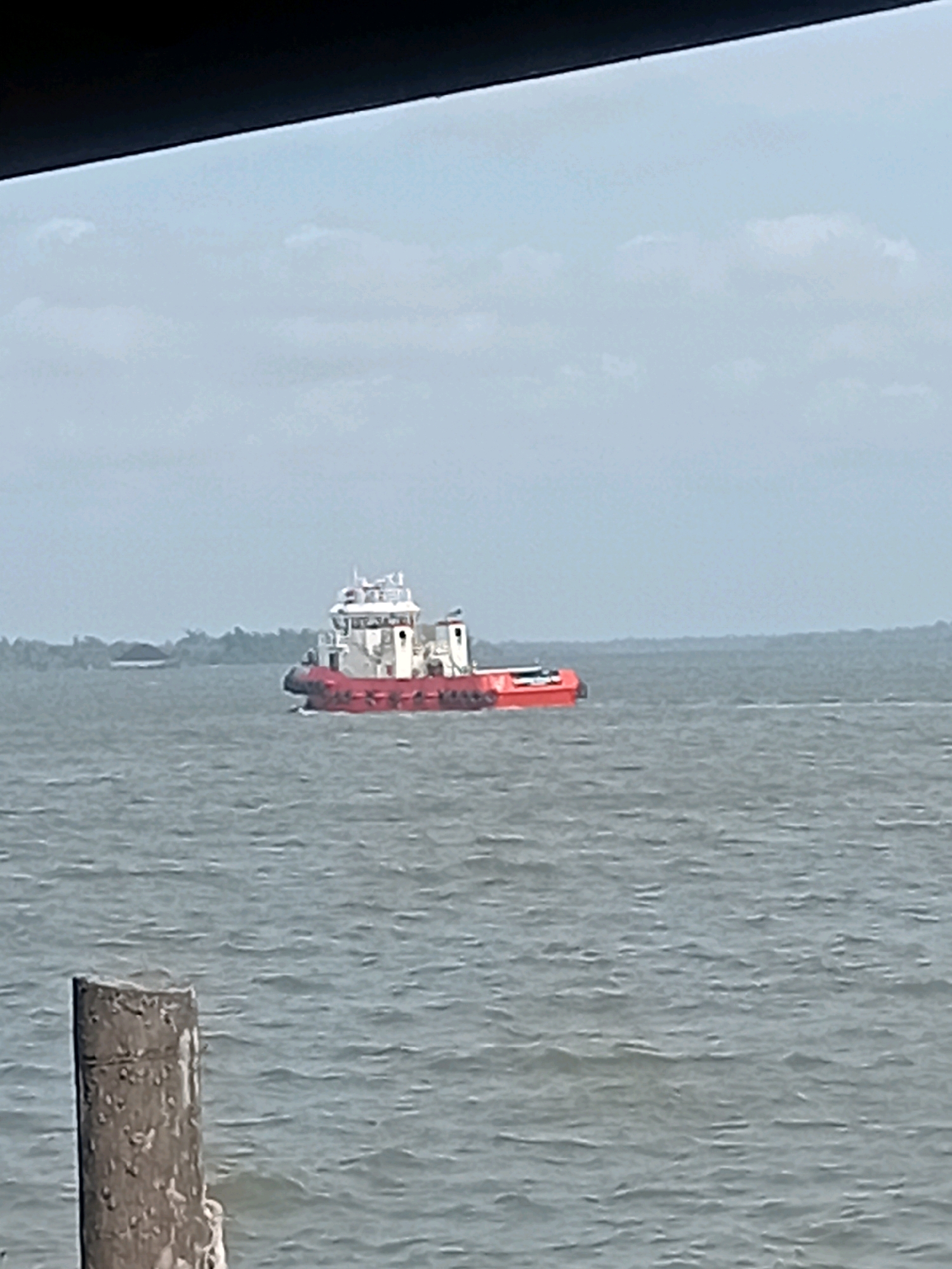A red and white tugboat is navigating on a body of water, likely a river or strait, given its proximity to land. The boat has a prominent white superstructure with multiple windows and a tall mast or antenna. The hull of the boat is a bright red. In the distance, a tree-lined shore is visible with some indistinct structures on it. In the foreground, a weathered wooden post, partially submerged in the water, stands vertically on the left side of the frame. The sky is overcast with scattered clouds. The water is choppy, indicating some movement or wind. The scene appears to be in an outdoor, coastal or riverine environment.