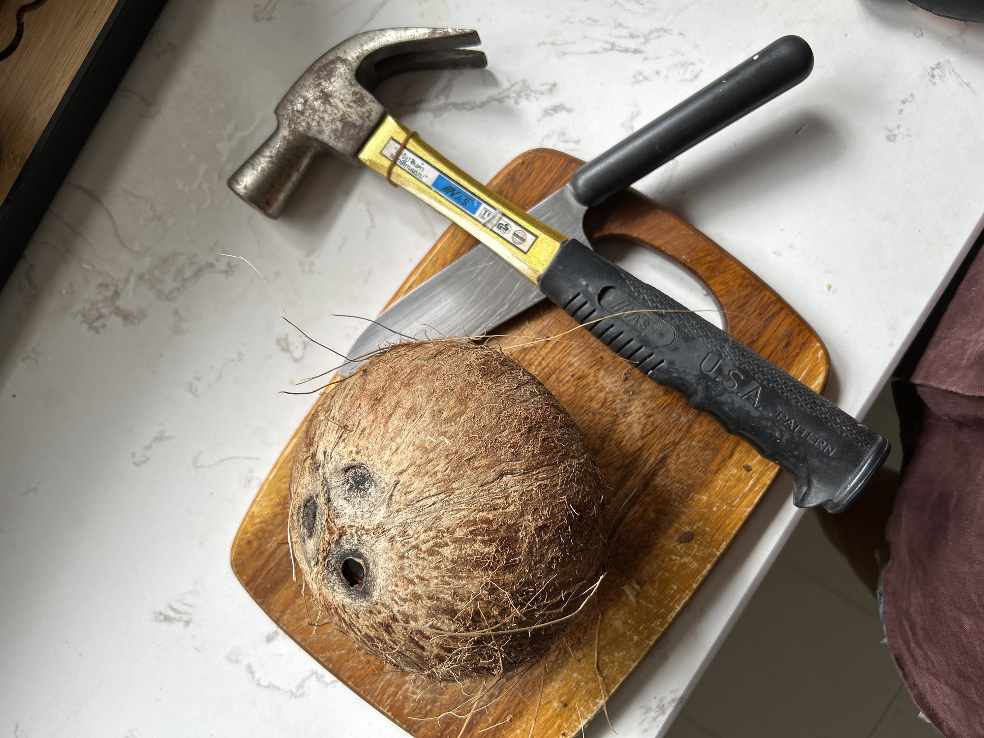 A whole coconut sits on a wooden cutting board. Beside it, a kitchen knife and a claw hammer are laid out. The background is a marble countertop. The image appears to be taken indoors, on a flat surface, possibly a kitchen or dining area. The lighting suggests it may be daytime, with artificial light contributing to the overall illumination. The scene is static, with no apparent activity or interaction between the objects. There are no people visible in the frame. The setting does not provide any specific indicators of its location within Chang Phueak, Thailand.