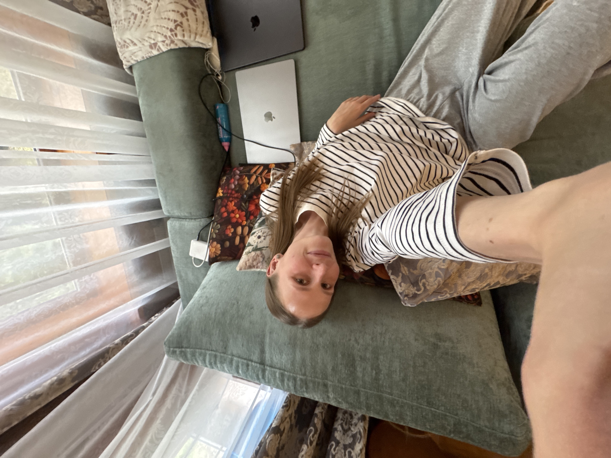 The image shows a woman lying on a green couch in what appears to be an indoor setting. She is wearing a striped shirt and grey pants. Two laptops, one dark and one light, are visible near her head. A window with white curtains is to her left. The location is Tomilino, Russia.