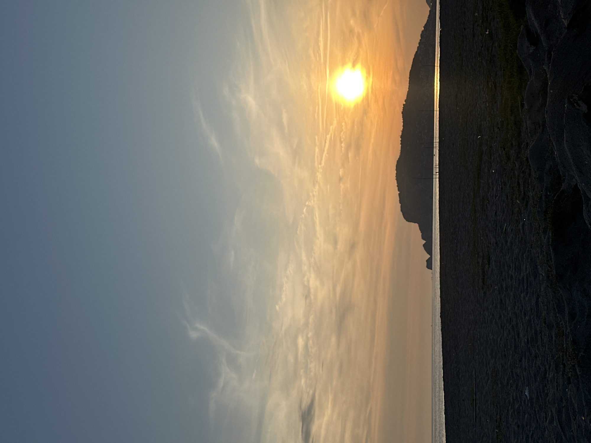 Captured at sunset in Alanya, Turkey, the image presents a serene landscape. The sky, a gradient of blue to orange, displays wispy clouds and aircraft contrails near the setting sun. A dark, mountainous silhouette stretches along the horizon, adjacent to a shoreline with a narrow strip of calm water. The foreground reveals a dark, sandy beach with visible texture and subtle gradations in tone. No people are visible.