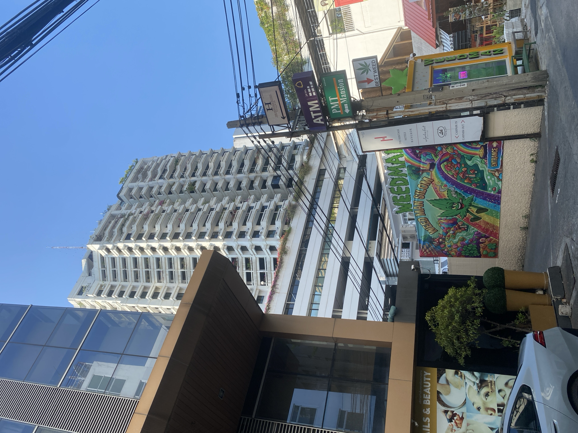The scene is an urban streetscape in Bangkok, Thailand, dominated by a tall white building with repetitive balconies and greenery. The image is taken from a low angle, showcasing the building's height. A modern brown and glass structure is prominent in the foreground. Power lines crisscross the sky. Street signs and advertisements line the buildings, including "The Heritage Hotels Bangkok," an "ATM," "PMT Mansion," and a large colorful sign advertising "WEEDMAN SILOM." A smaller sign reads "BEST WEED." Another advertises "NAILS & BEAUTY." A car is partially visible on the right. The overall environment suggests a bustling commercial area.