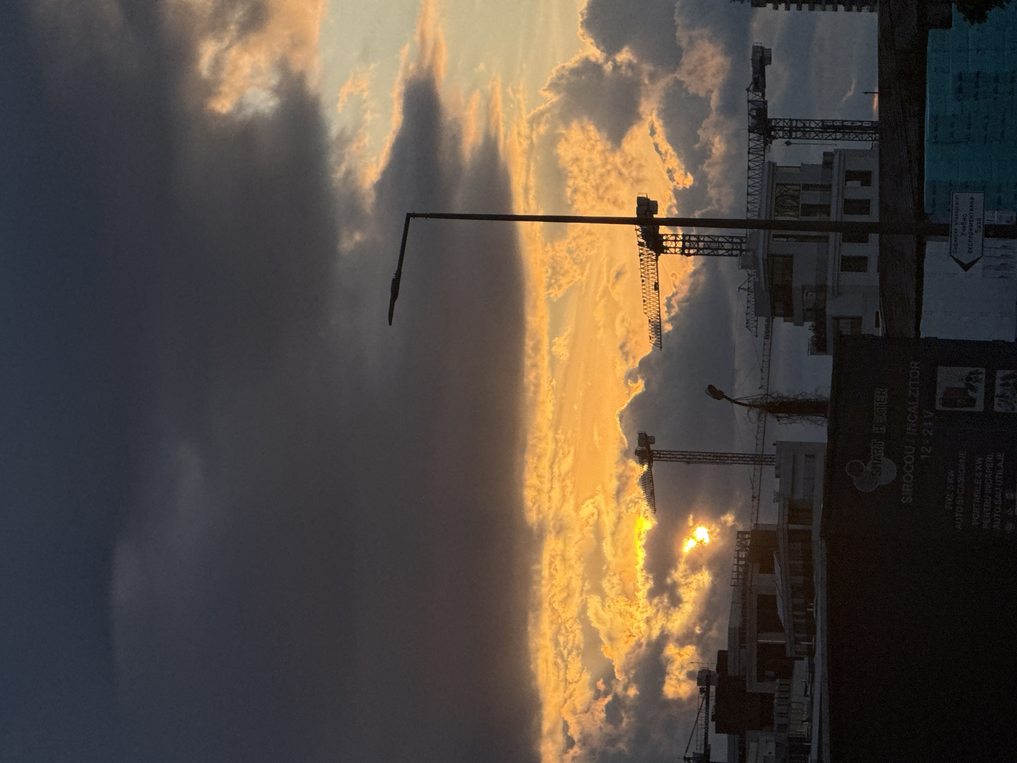 This is a vertically oriented photograph taken at sunset in Sofia, Bulgaria. The sky is dramatically lit with a vibrant orange and yellow glow from the setting sun, which is partially obscured by dramatic, dark clouds that cast long shadows. Silhouetted against the colorful sky are several construction cranes, indicating an active construction site. In the background, buildings are visible, also in silhouette.
The lower portion of the image features a dark, vertical sign with text. The visible text includes "SHARITHEATER", "SIROCOU/INCALZITOR 12-24V", and also some text in Cyrillic which translates to "Sofia University, Educational Experimental Base". This suggests the location is likely near an educational or research facility related to Sofia University. The presence of the sun setting behind clouds and the silhouettes of construction cranes create a somewhat dramatic and potentially moody atmosphere. The time of day is clearly evening, as indicated by the sunset.